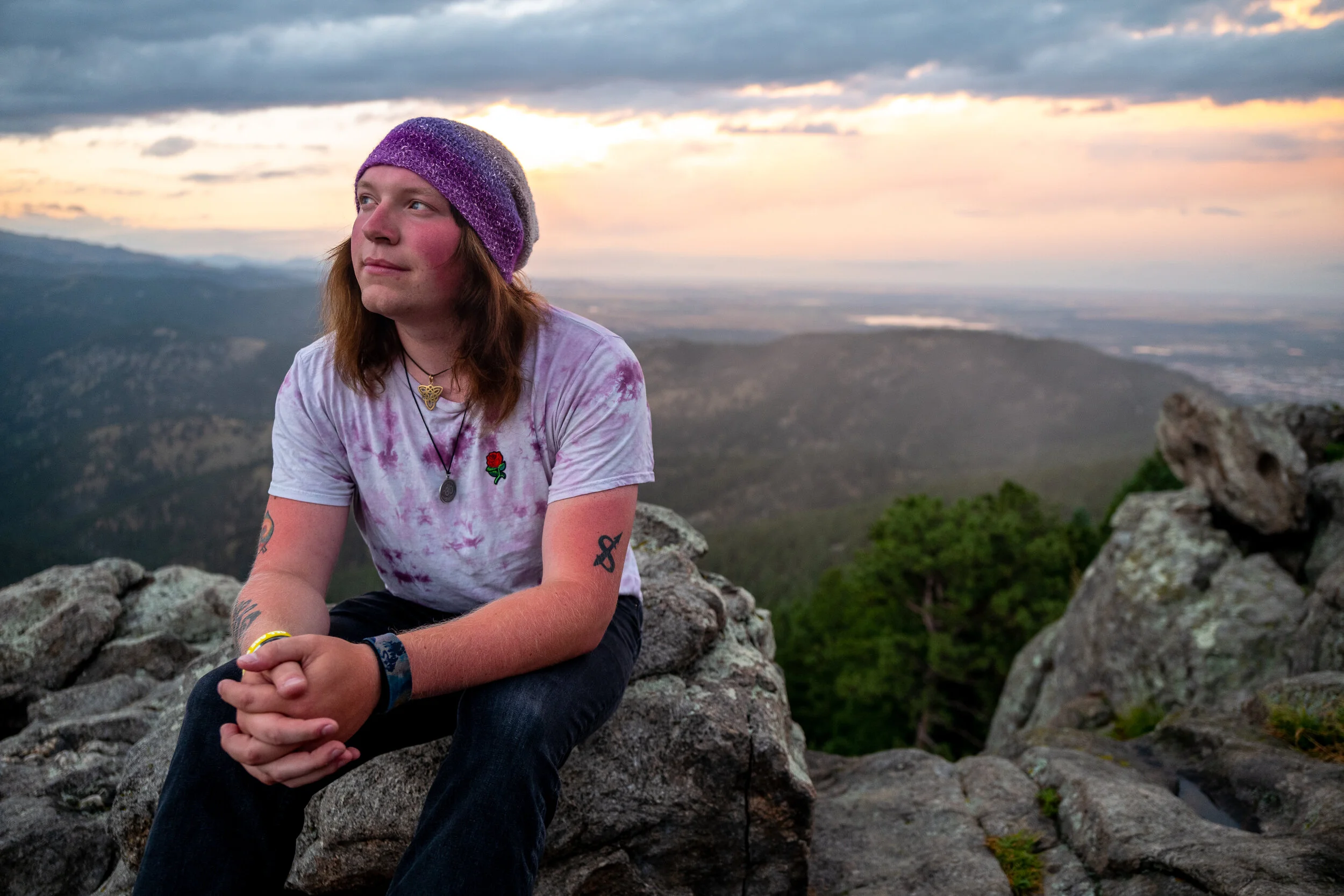 A person with shoulder-length brown hair wearing a purple knit hat, a tie-dye purple and white t-shirt with a red rose patch, dark jeans, and layered necklaces, sitting on a rock atop a mountain with a scenic valley, trees, and a sunset sky in the ba