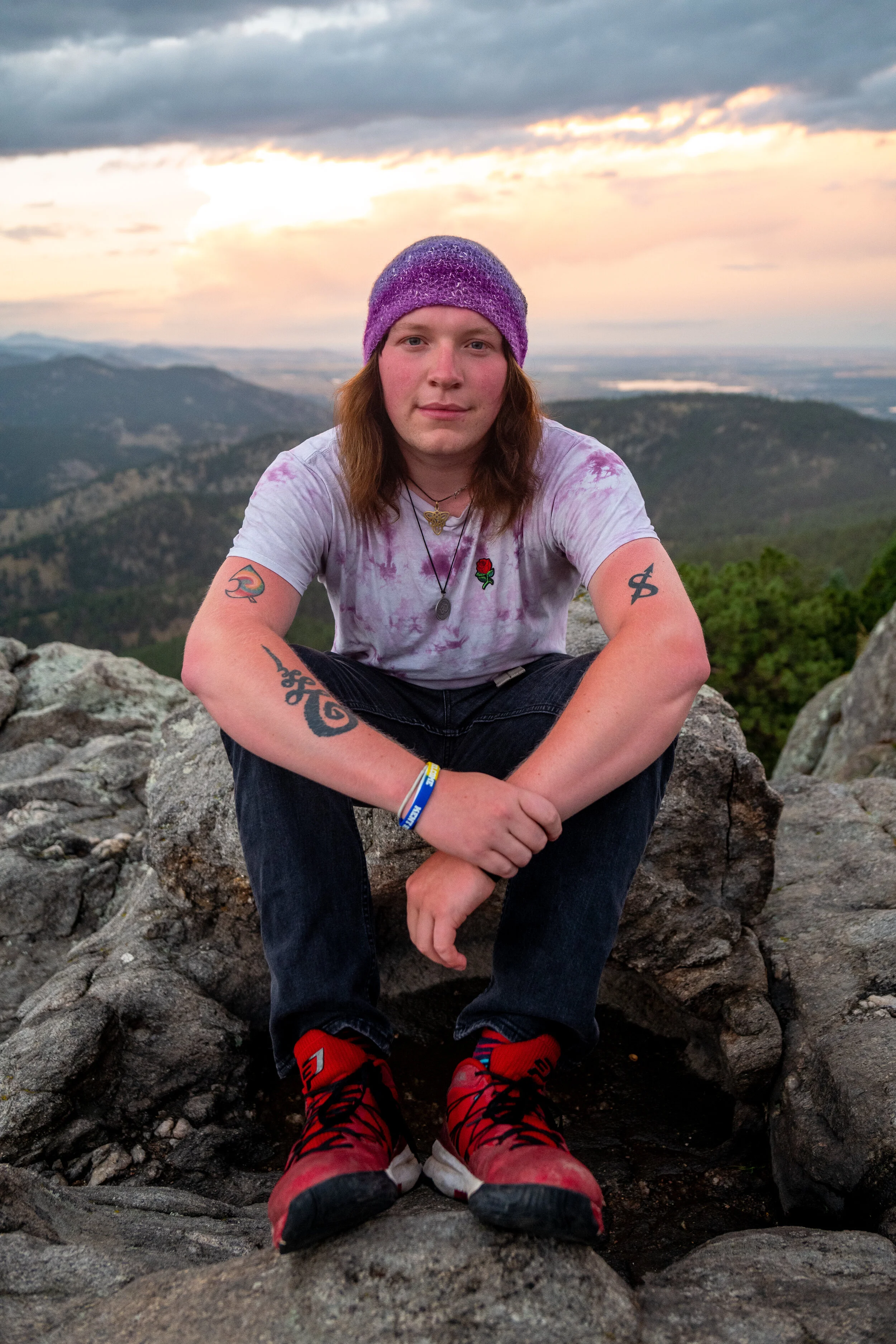 A person with long hair, wearing a purple knit hat, tie-dye shirt, black pants, and red hiking shoes, sitting on a rock with a mountain and sunset in the background.