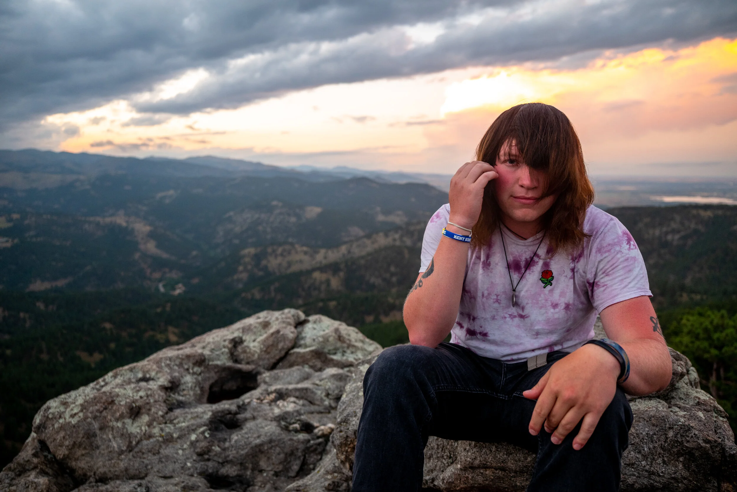 A young woman with brown hair sits on a large rock on a mountain, with a scenic view of rolling hills and a cloudy sky at sunset in the background.