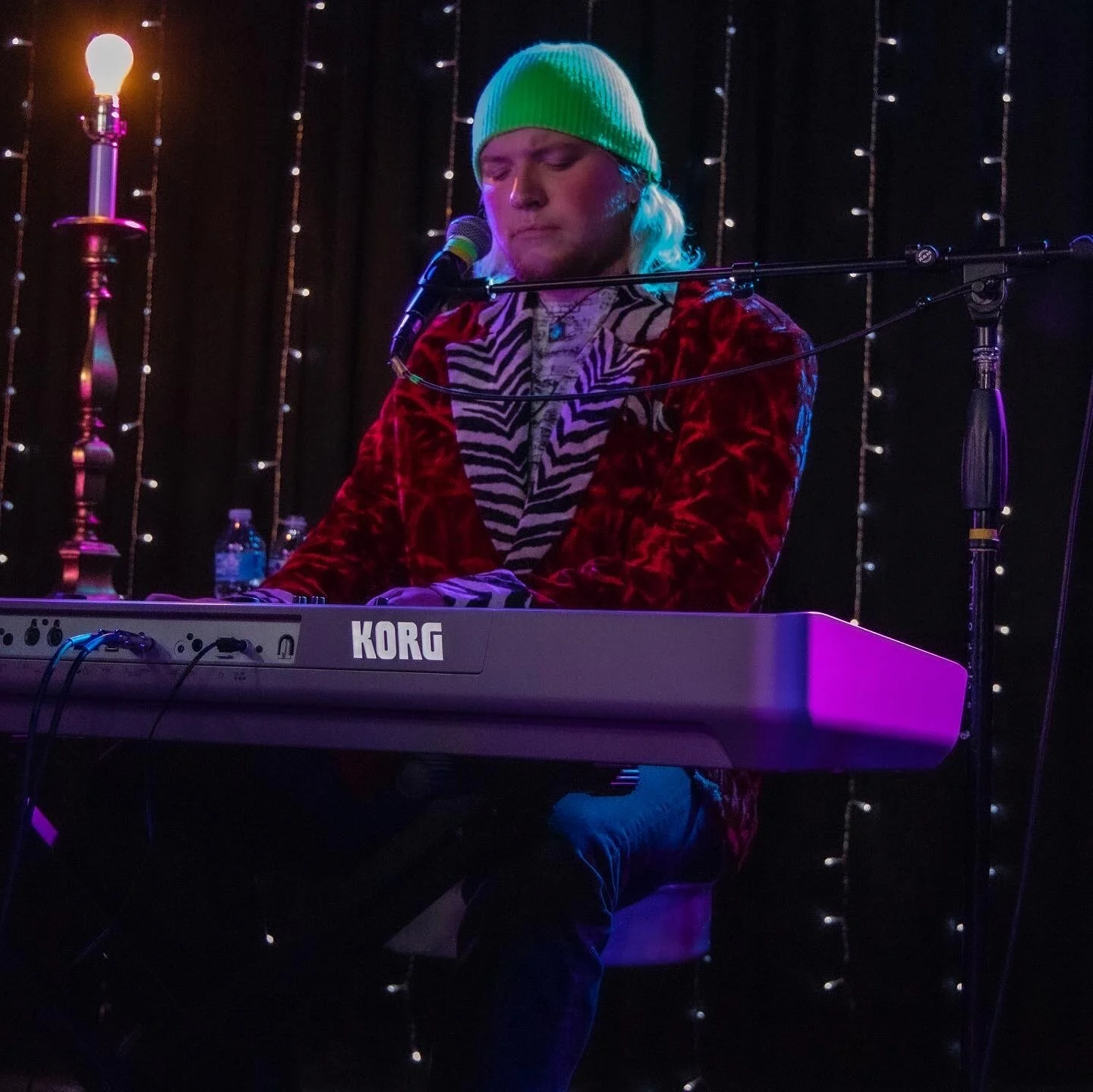 Person with long blond hair wearing a green beanie and a red velvet jacket playing a Korg keyboard on stage with string lights in the background.