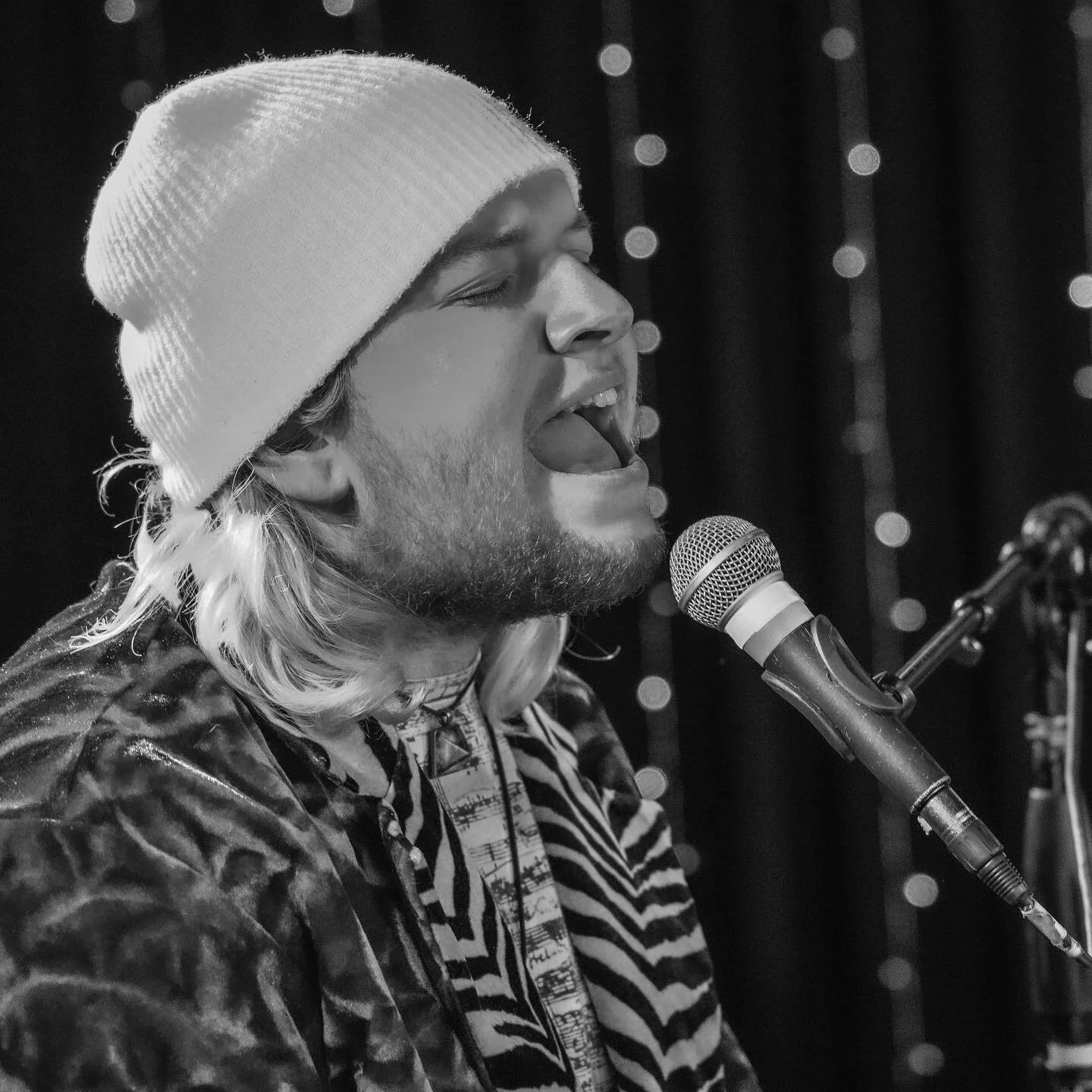 A man singing passionately into a microphone during a performance, wearing a beanie hat and a patterned jacket.