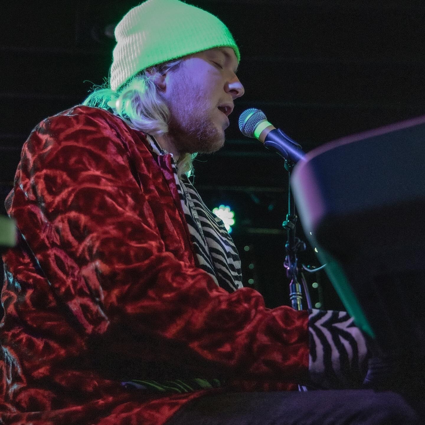 A musician singing into a microphone on stage, wearing a neon green beanie, a red velvet jacket, and a zebra-striped shirt.