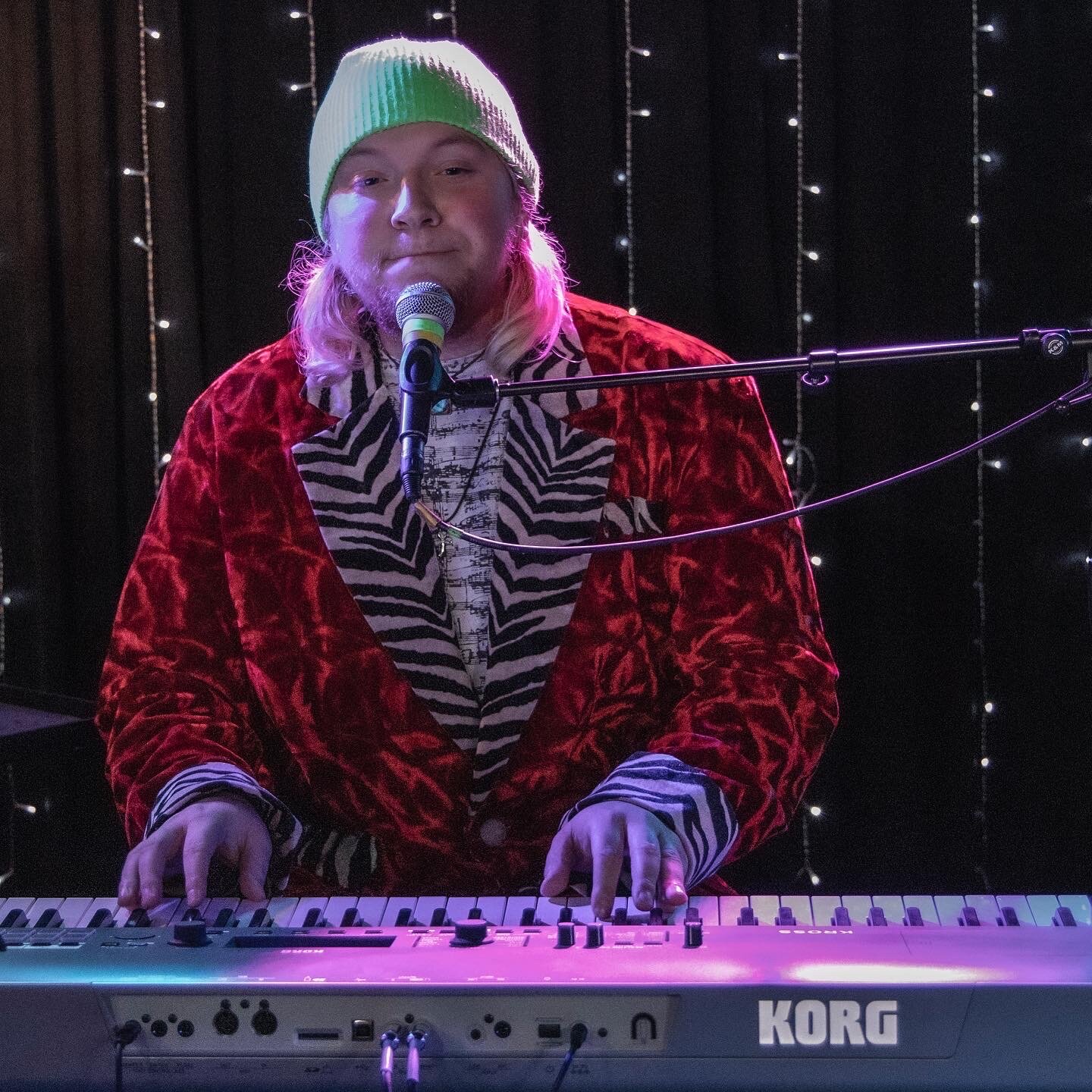 A person with long blond hair wearing a lime green beanie, a red velvet jacket, and a zebra-patterned shirt, plays a keyboard and sings into a microphone on a stage decorated with string lights.