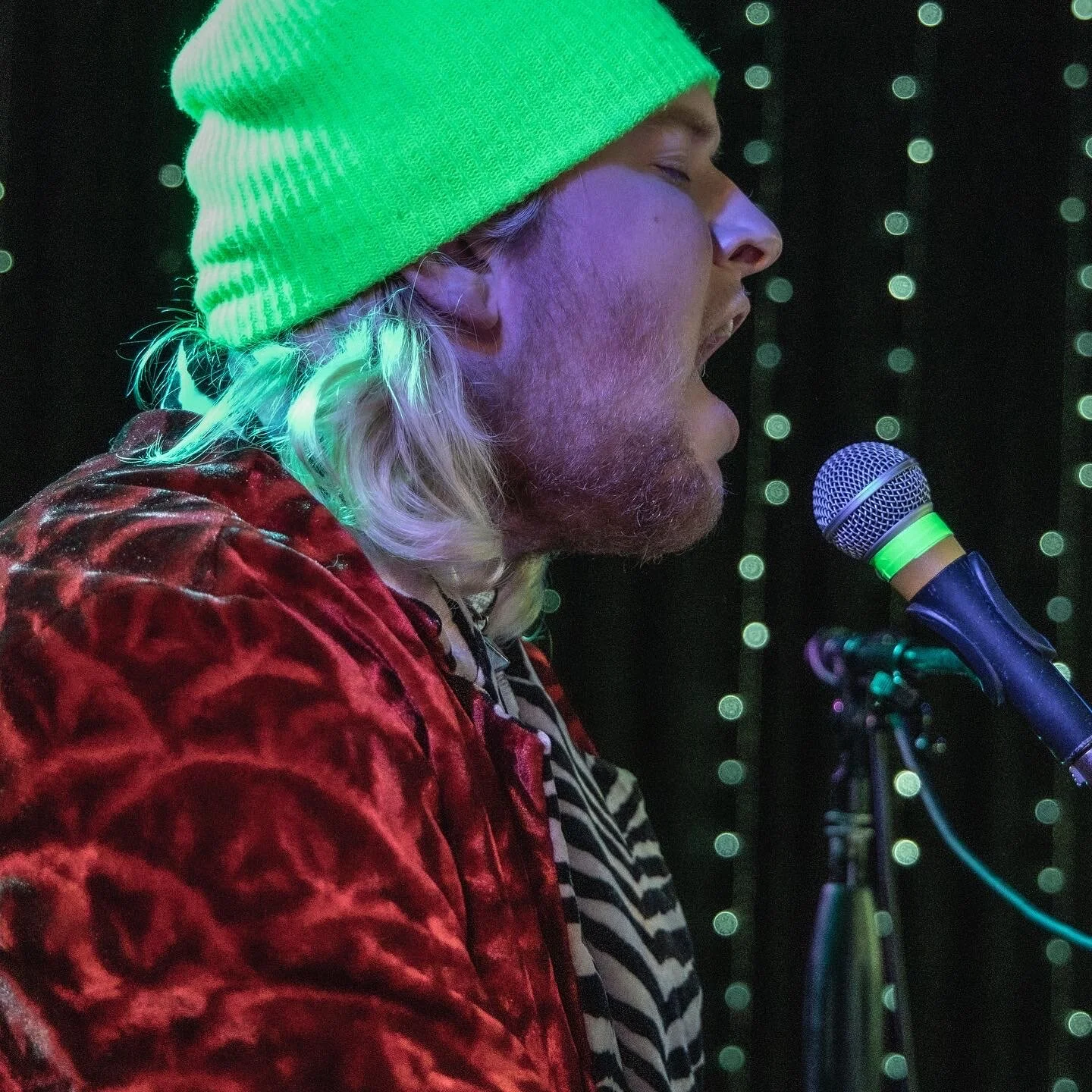 Performing musician singing into a microphone on stage, wearing a bright green beanie, red velvet jacket, and striped shirt, with green LED lighting in the background.