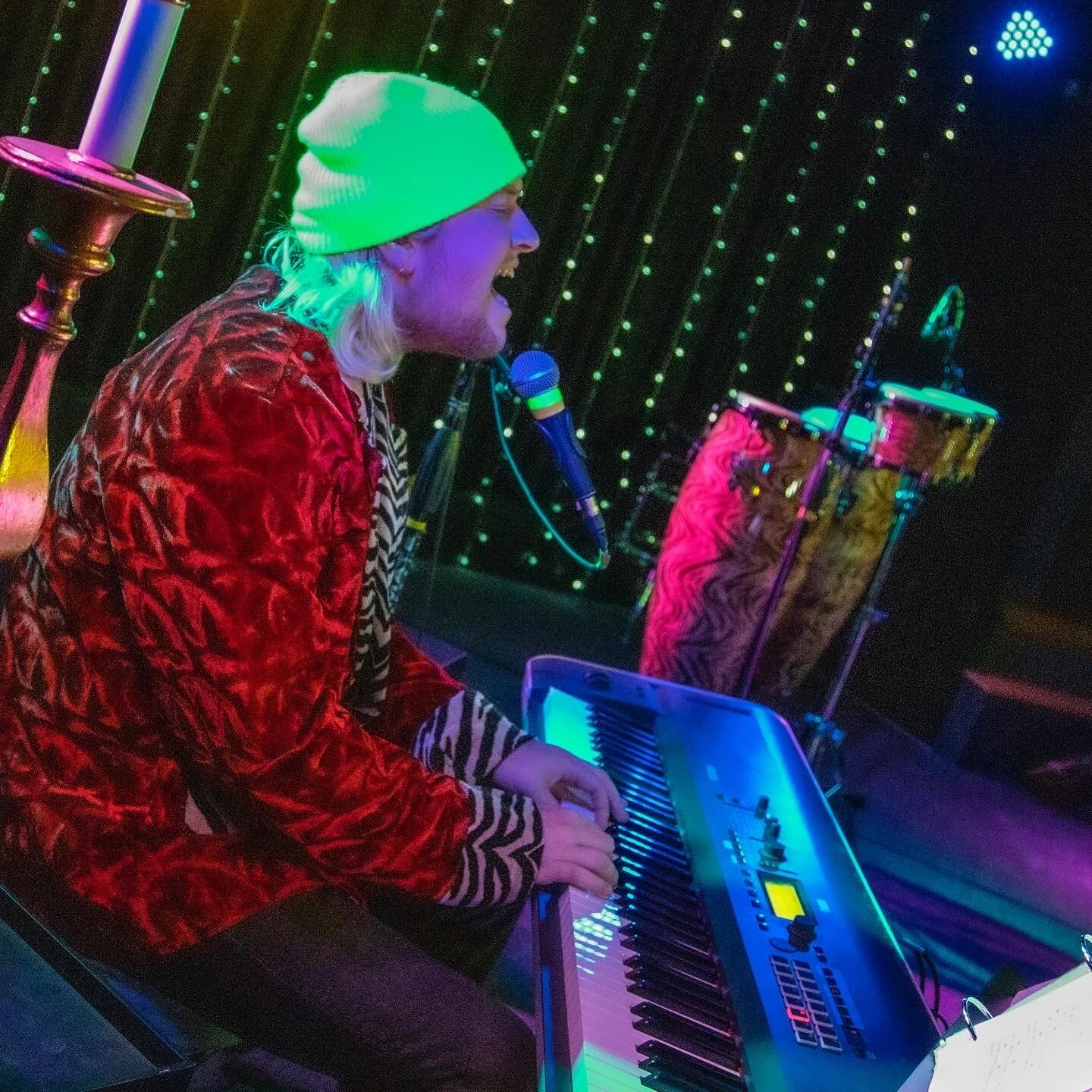 A man with long, blonde hair and a beard, wearing a white beanie and a red velvet jacket, is playing a keyboard on stage, singing into a microphone with a colorful, neon-lit background.
