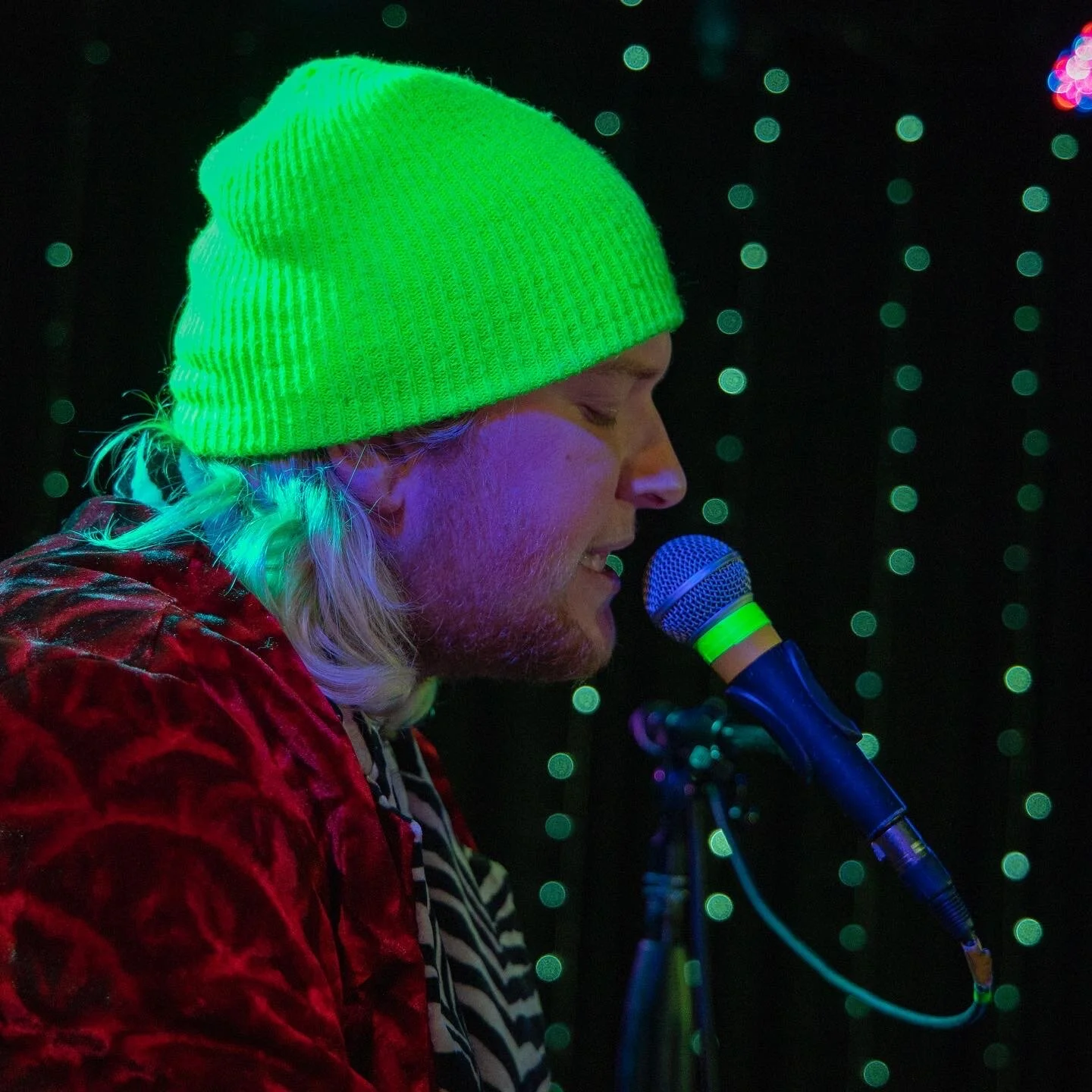 A man with long blond hair, wearing a bright green beanie and a red velvet jacket, singing or speaking into a microphone on stage with a dark background and green decorative lights.