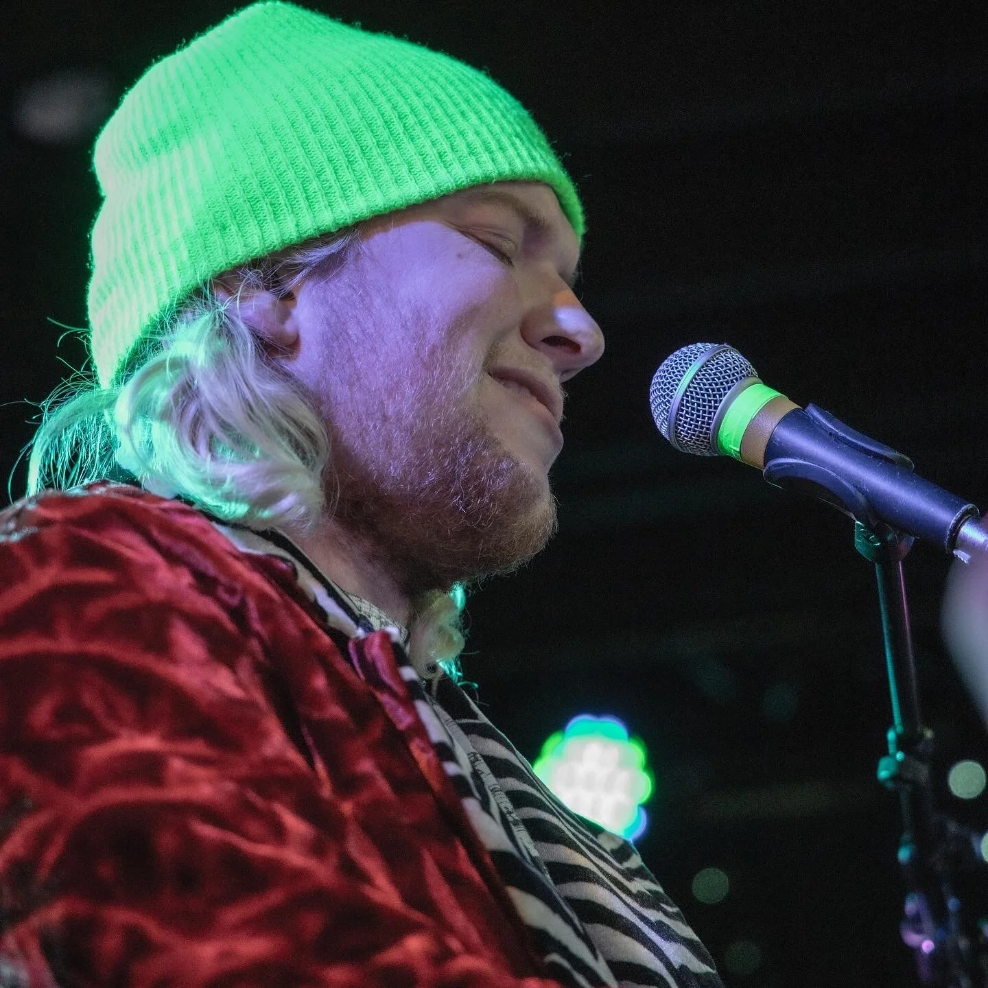 A man with long blond hair, a beard, wearing a green knit cap, singing or speaking into a microphone on stage with colorful lights in the background.