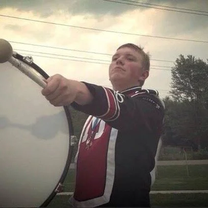 A young man wearing a black and red jacket holding a drumstick, standing outdoors with trees and power lines in the background.
