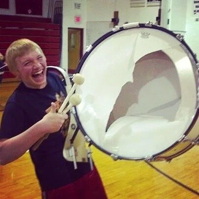 A boy with blonde hair smiling happily while holding a large bass drum and drumsticks, in a gymnasium or auditorium setting.