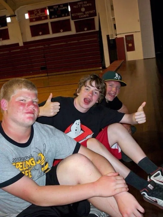 Three boys sitting on the gym floor inside a gymnasium, smiling and giving thumbs up. The boy on the left has a red face, short blonde hair, and is wearing a gray T-shirt. The boy in the middle has curly hair, is wearing a black Ghostbusters T-shirt,