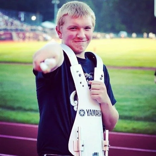 A young male track athlete on a sports field, pointing toward the camera with a smile, wearing a black shirt and a white race bib with Yamaha branding, likely during a track event.