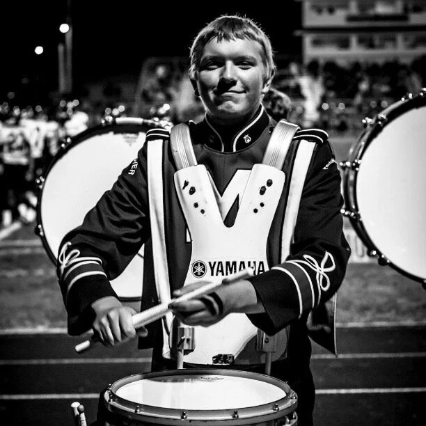 A young man in a marching band uniform is holding a drumstick, standing in front of a row of bass drums, at night.