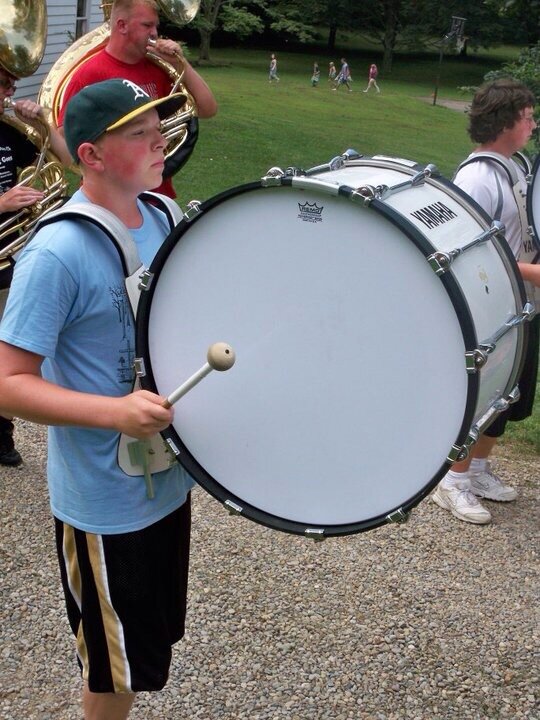 Young boy playing bass drum in a marching band outdoors, with other band members and people walking in the background.