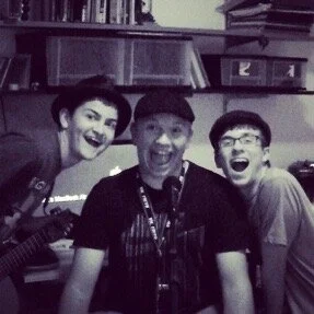 Three smiling people posing together indoors with bookshelves in the background.