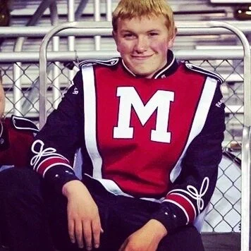 Young boy wearing a red and black football letterman jacket with a large white letter 'M' on the front, sitting in front of a chain-link fence.