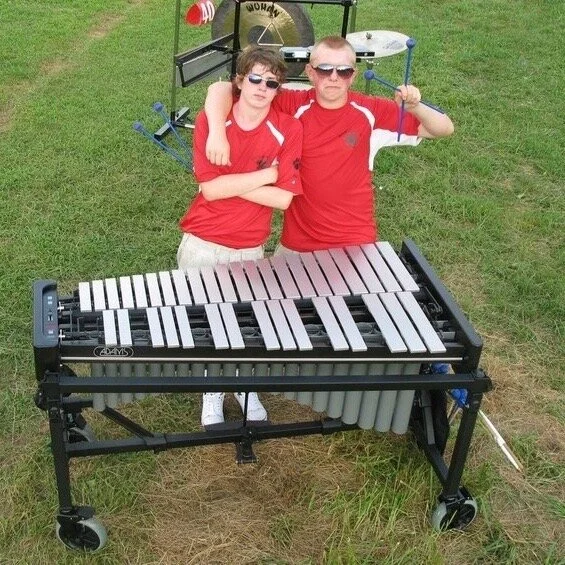 Two boys in red shirts standing outdoors on grass, with an electronic xylophone in front of them, and musical percussion instruments in the background.