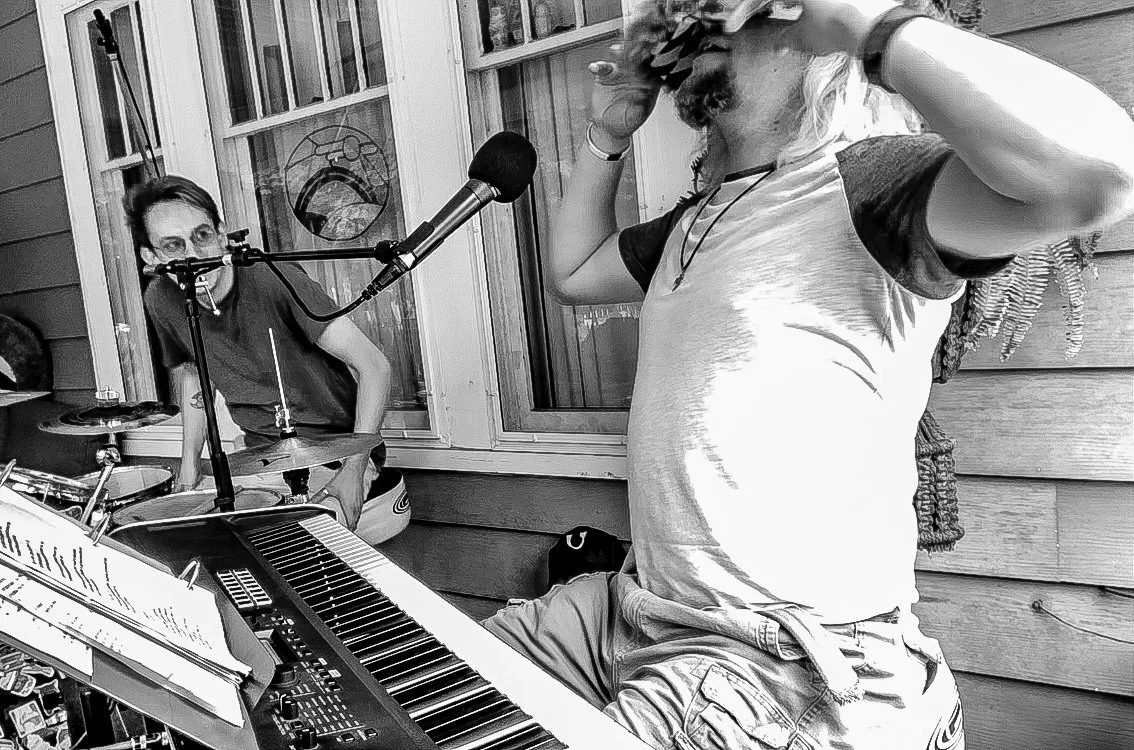 A musician sitting at a keyboard on a porch, with a woman singing into a microphone and playing drums nearby.