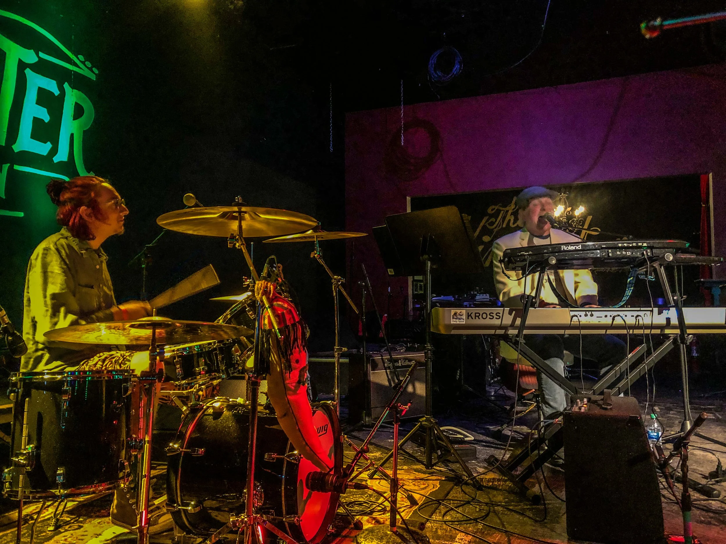 Musicians performing live on stage, one playing drums and the other singing and playing a keyboard, in a dimly lit venue with colorful lighting.