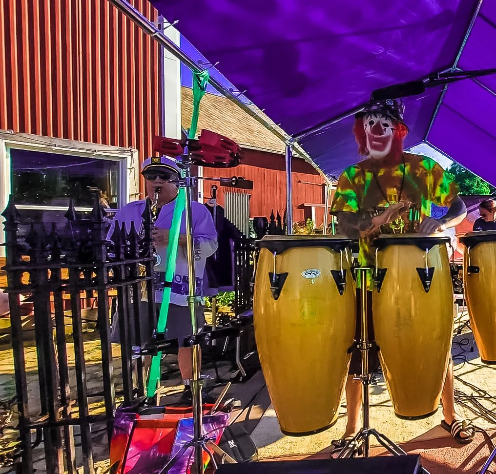 A person wearing a clown mask and colorful shirt playing drums, alongside another person playing a saxophone, under a purple canopy.
