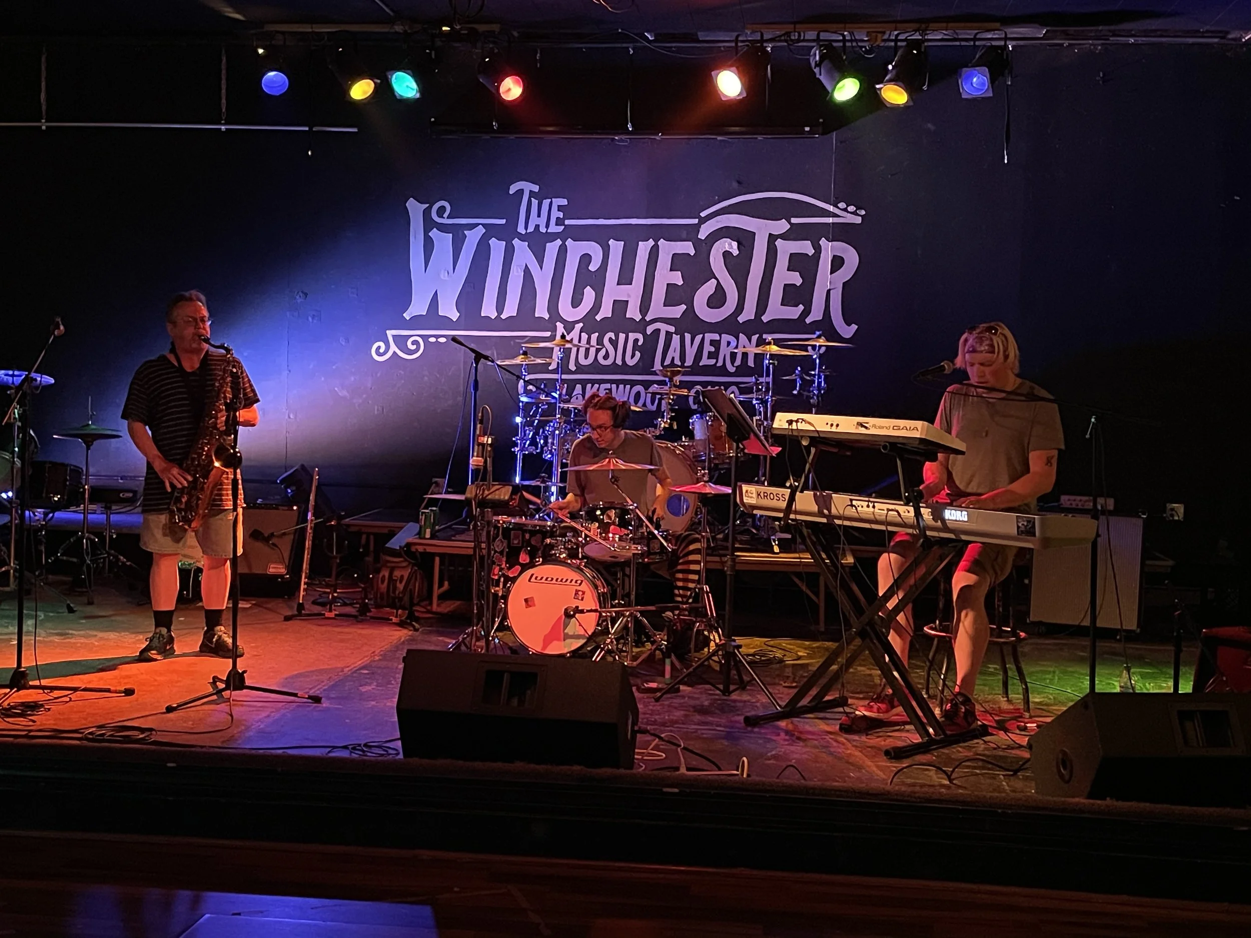 A band performing on stage at The Winchester Music Tavern, with a saxophonist, drummer, and keyboardist under colorful stage lighting.