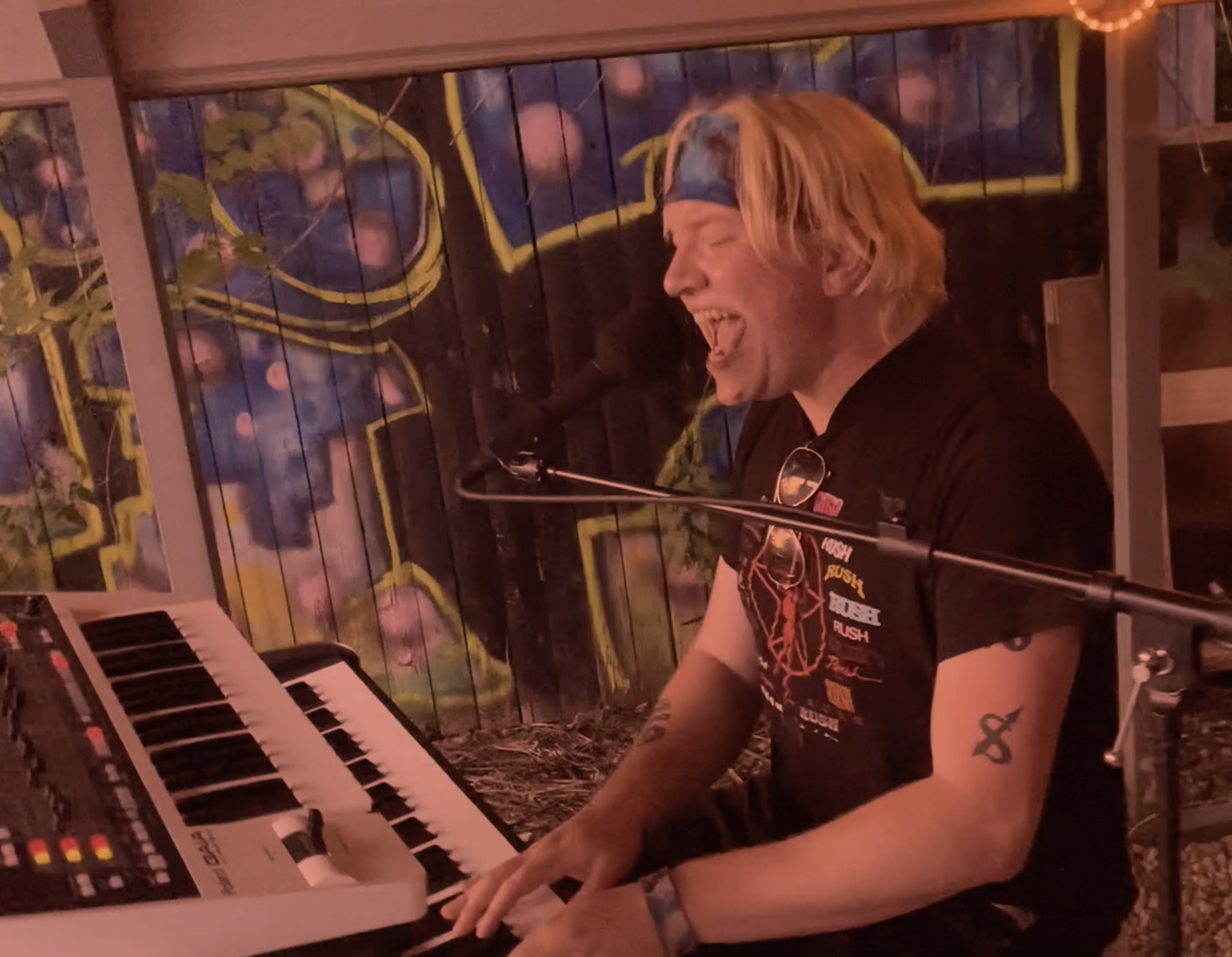 A man with blond hair and a blue headband singing passionately while playing a keyboard at a live music performance, with graffiti art on the wall behind him.