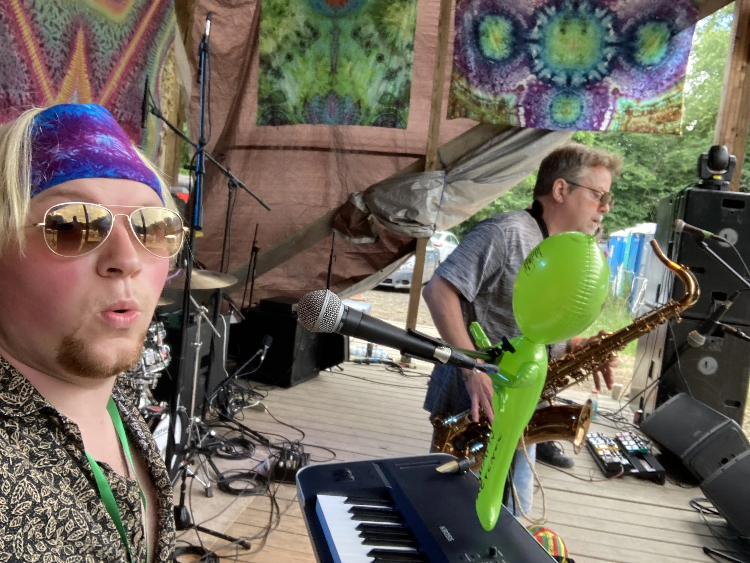 A musician with sunglasses and a colorful bandana singing into a microphone during an outdoor performance, with a man in the background playing a saxophone and tie-dye banners hanging behind them.