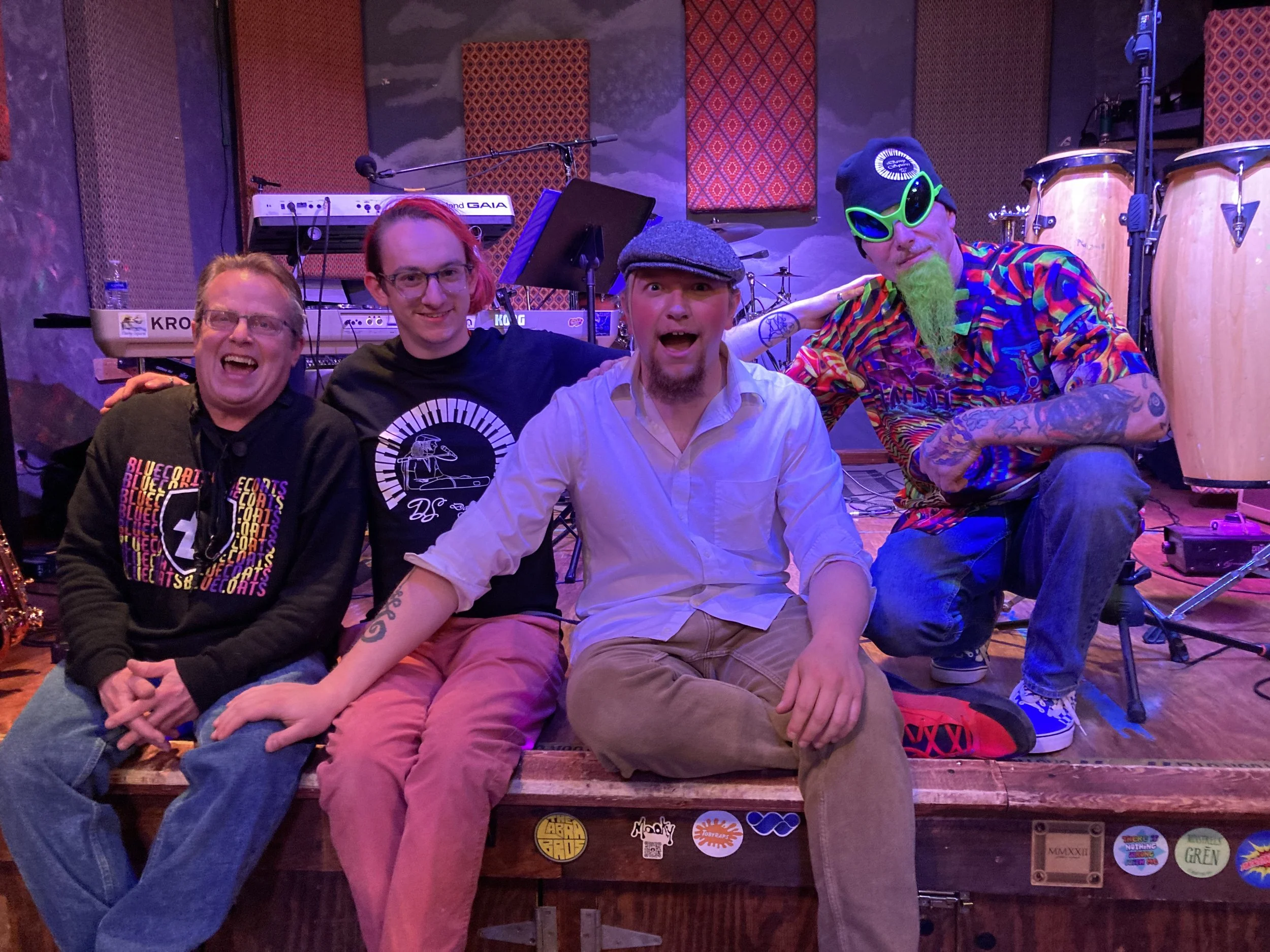 Four men sitting on a stage after a performance, smiling and posing for the photo, with musical instruments and equipment in the background.