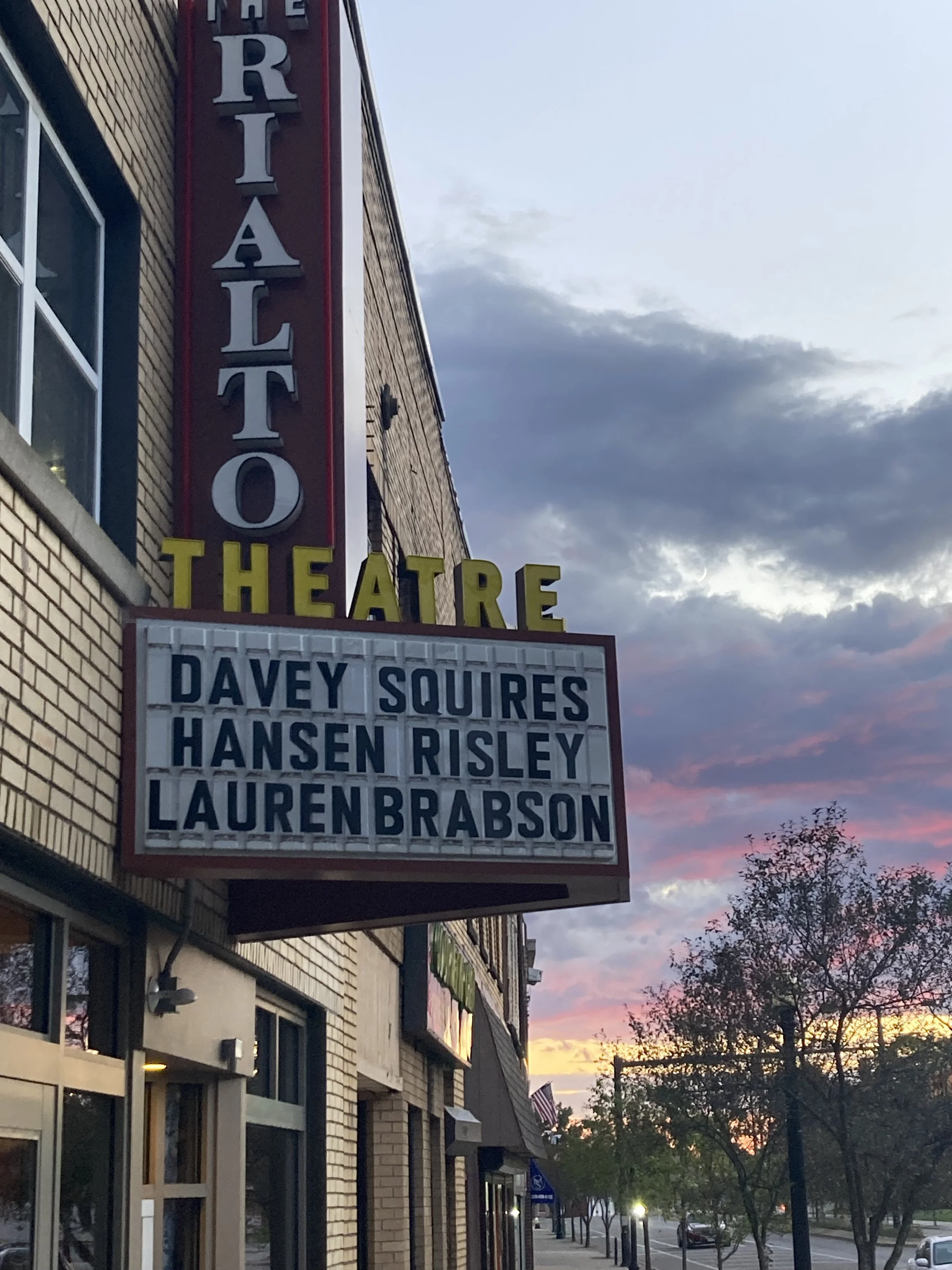 A theater marquee displaying the names of performers Davey Squires, Hansen Risley, and Lauren Brabson against a sunset sky with clouds and trees.