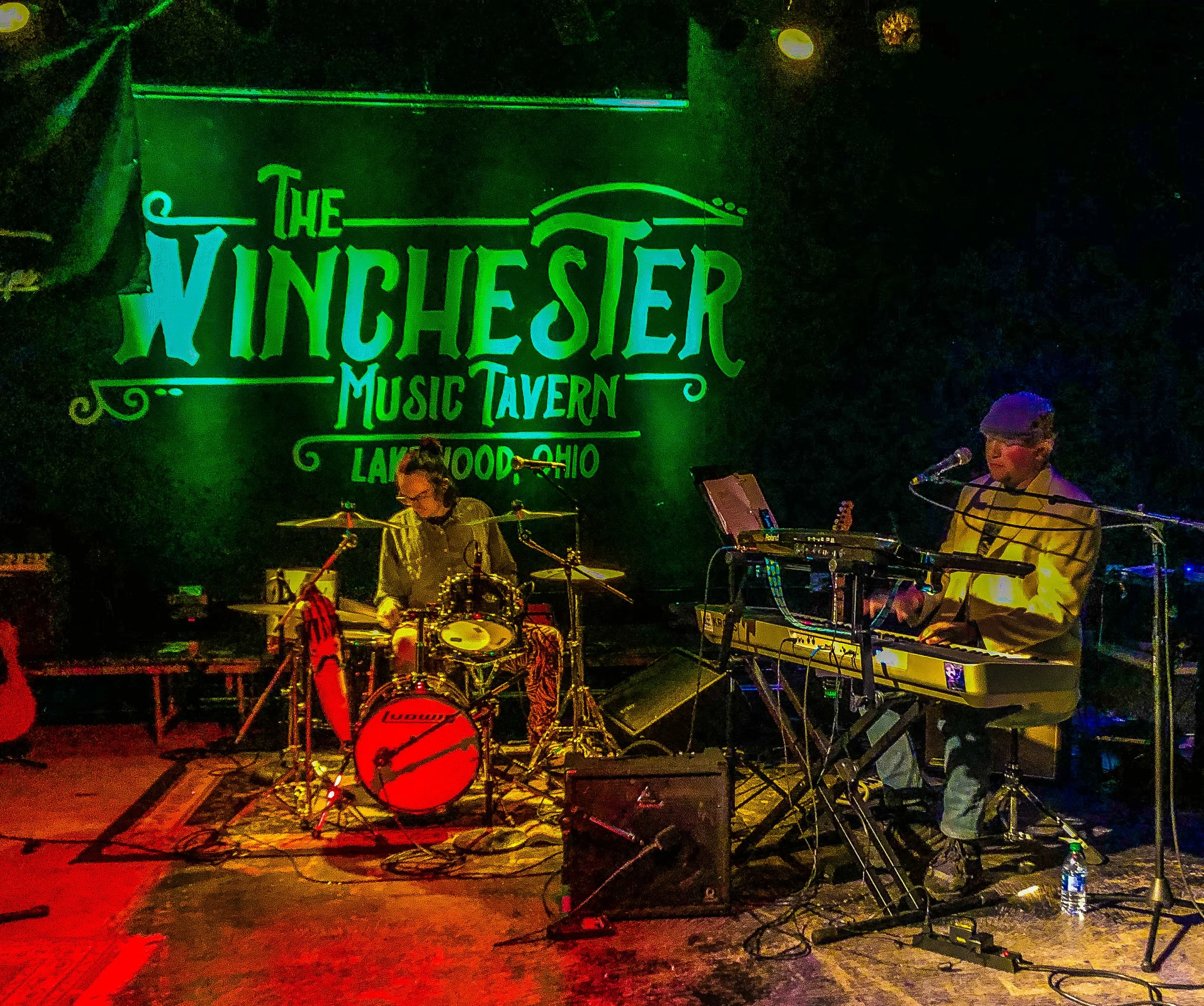 Two musicians performing on a stage at The Winchester Music Tavern in Lakewood, Ohio. One is playing drums, the other is on keyboard and singing into a microphone. The background features a large sign with the venue's name.