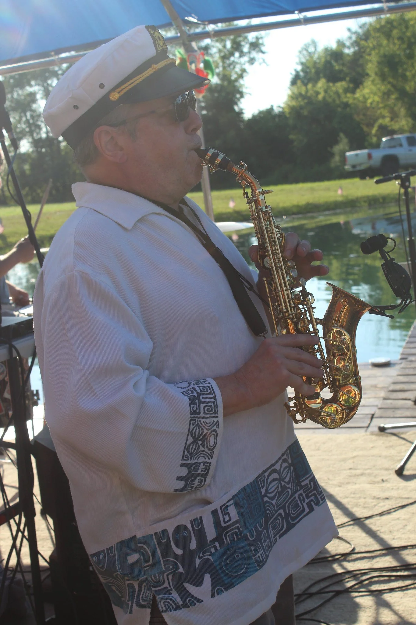 A man in a captain's hat and sunglasses playing a saxophone outdoors near a body of water, with trees and a truck in the background.