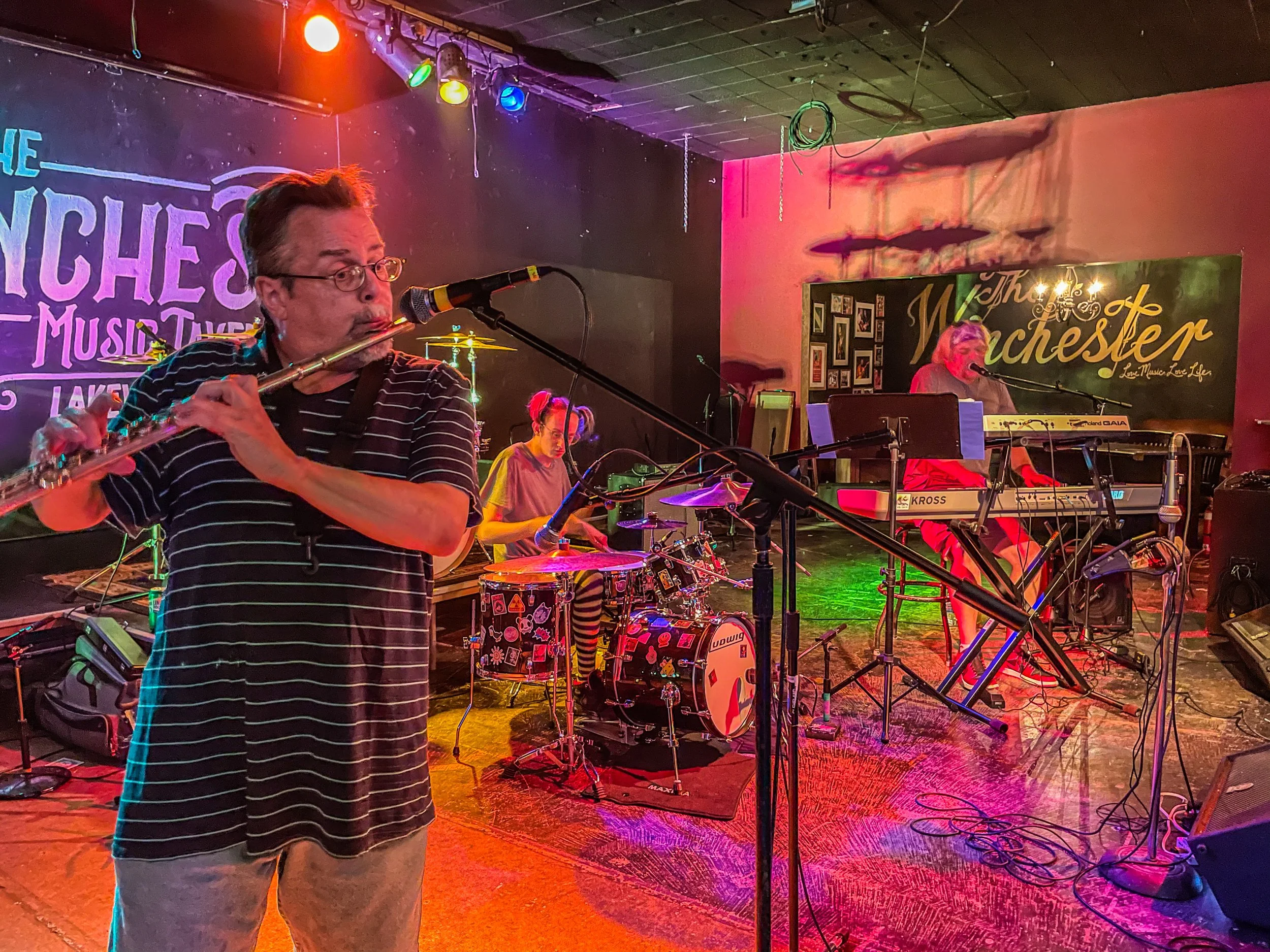 A band performing on stage in a music venue, featuring a man playing a flute, a drummer, and a woman at a keyboard, with colorful stage lighting and a large sign in the background that reads 'The Winchester'.
