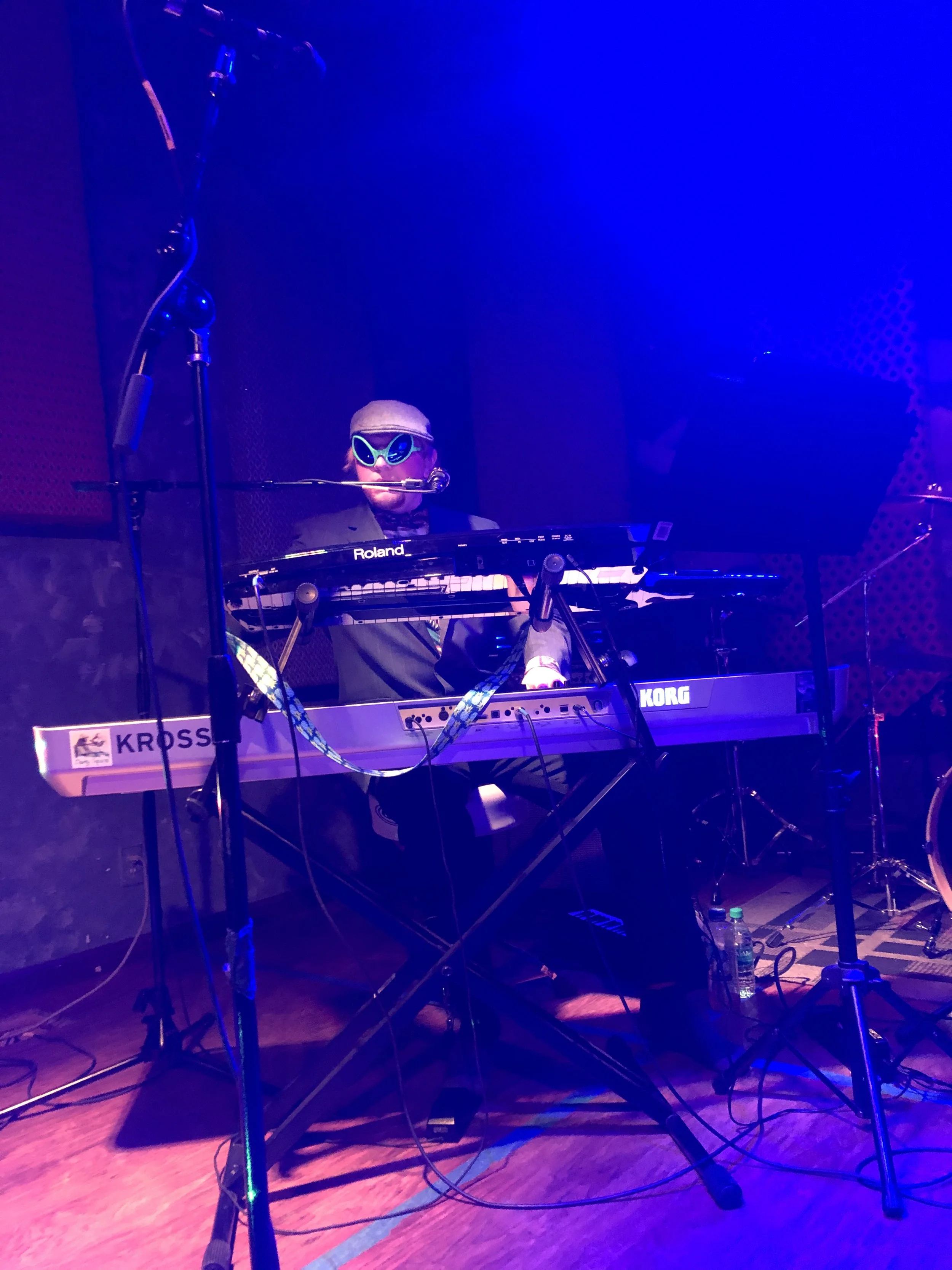Musician wearing sunglasses and a hat playing a keyboard on stage with blue lighting.