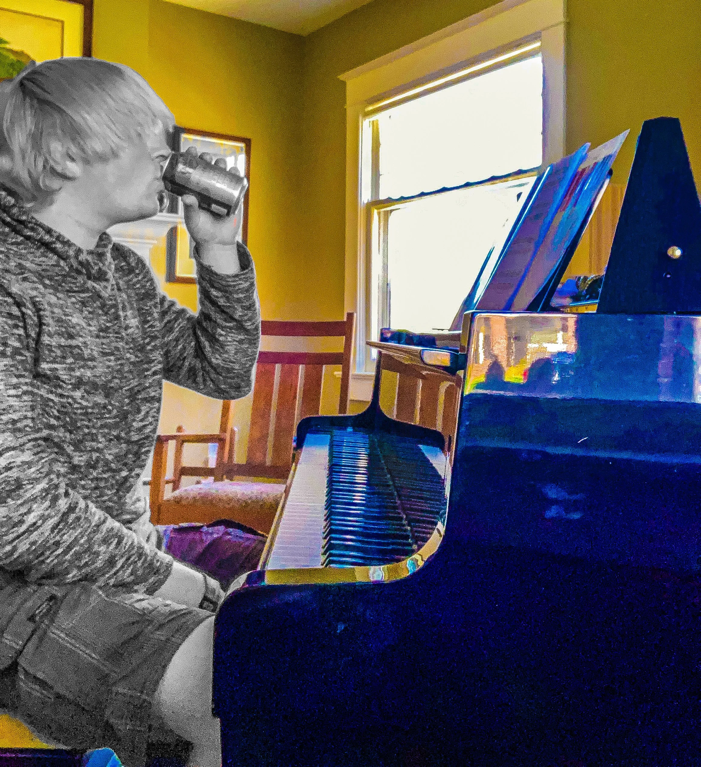 A young person sitting at a black grand piano, drinking from a can, wearing a hoodie and shorts, in a room with a yellow wall and a window.