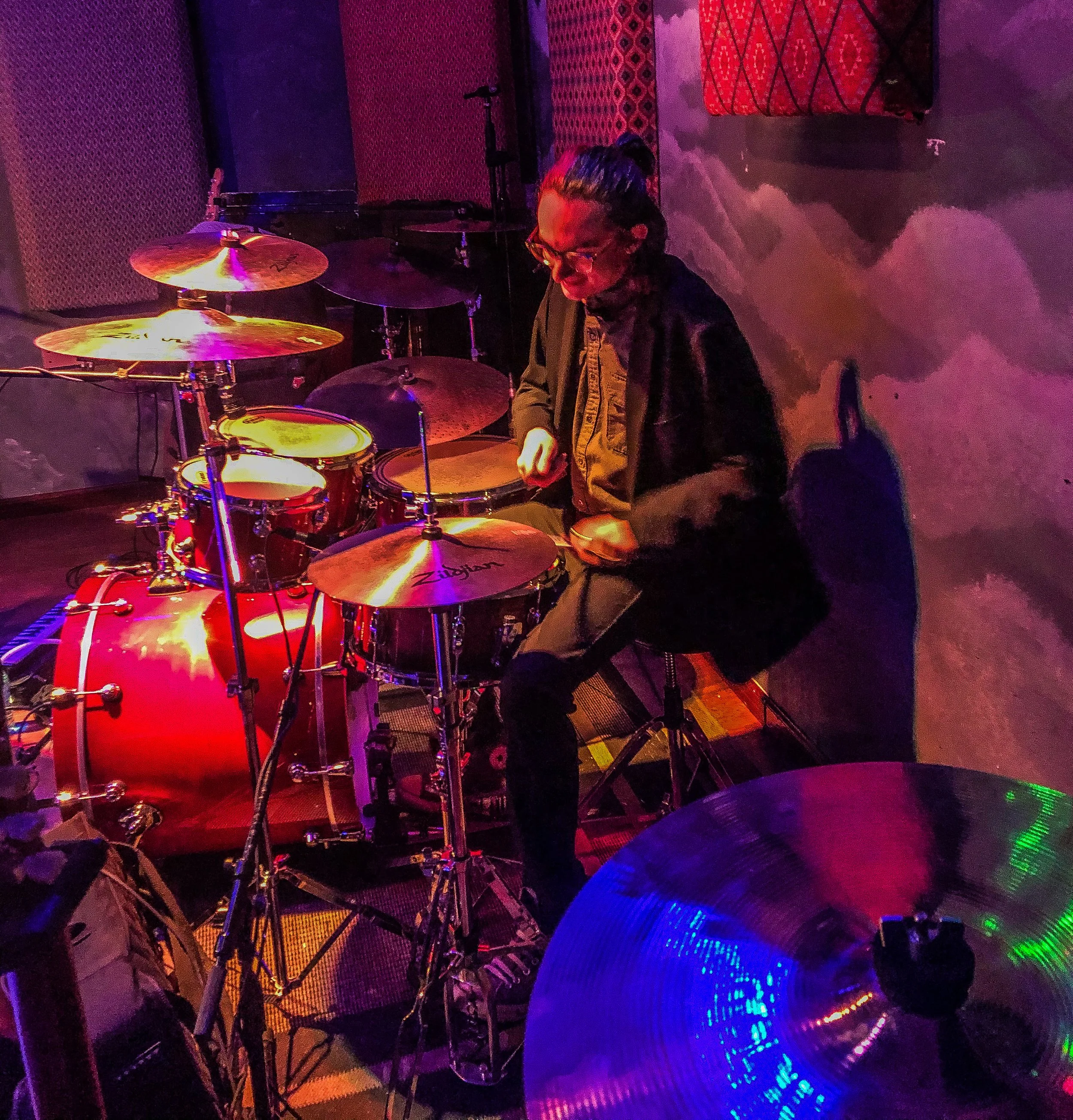 A person playing drums in a dimly lit room with colorful lights reflecting off the cymbals and drums.