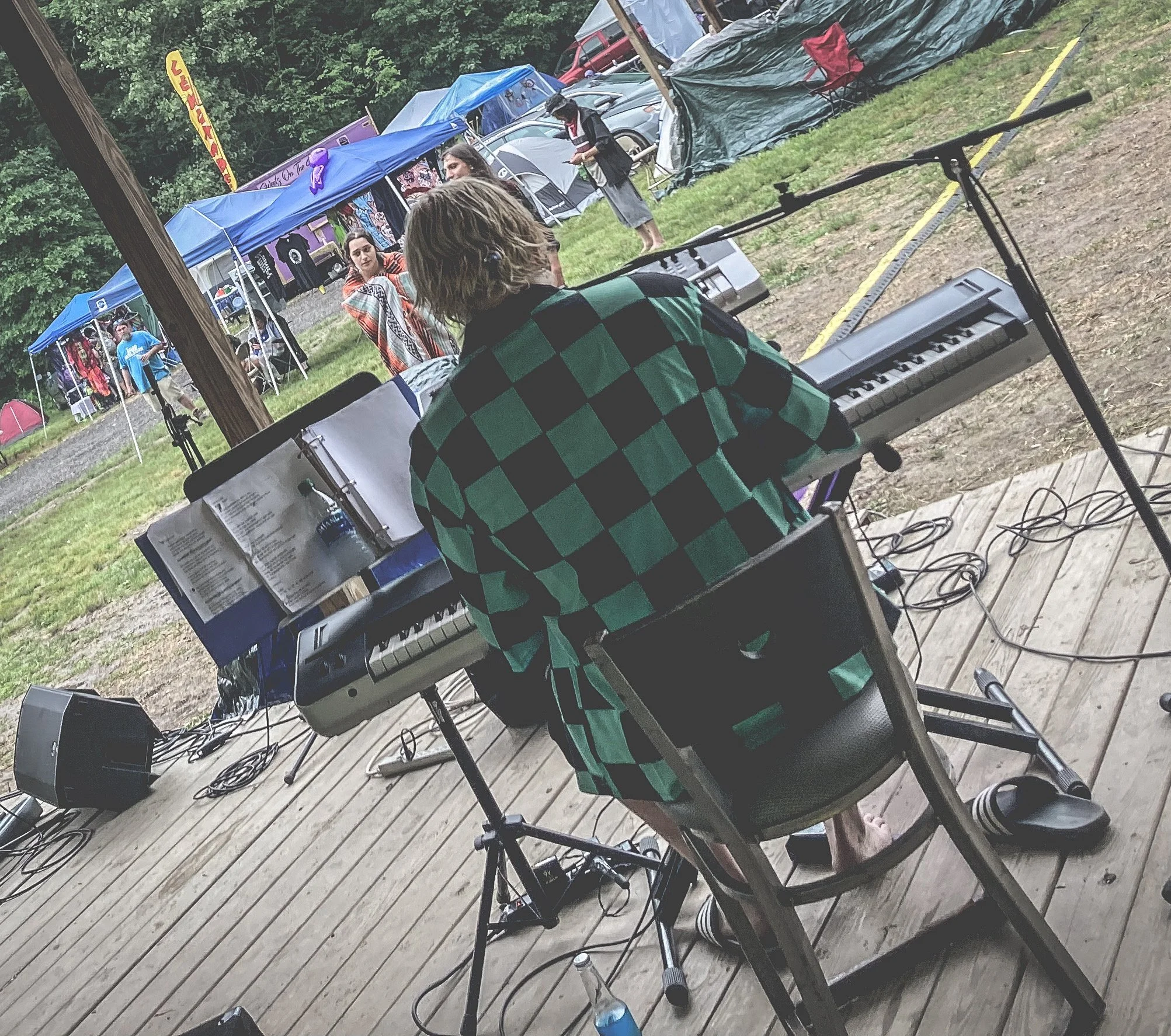 A musician with blonde hair and a black and green checkered shirt playing a keyboard on an outdoor stage at a festival. Tents and festival attendees are visible in the background.
