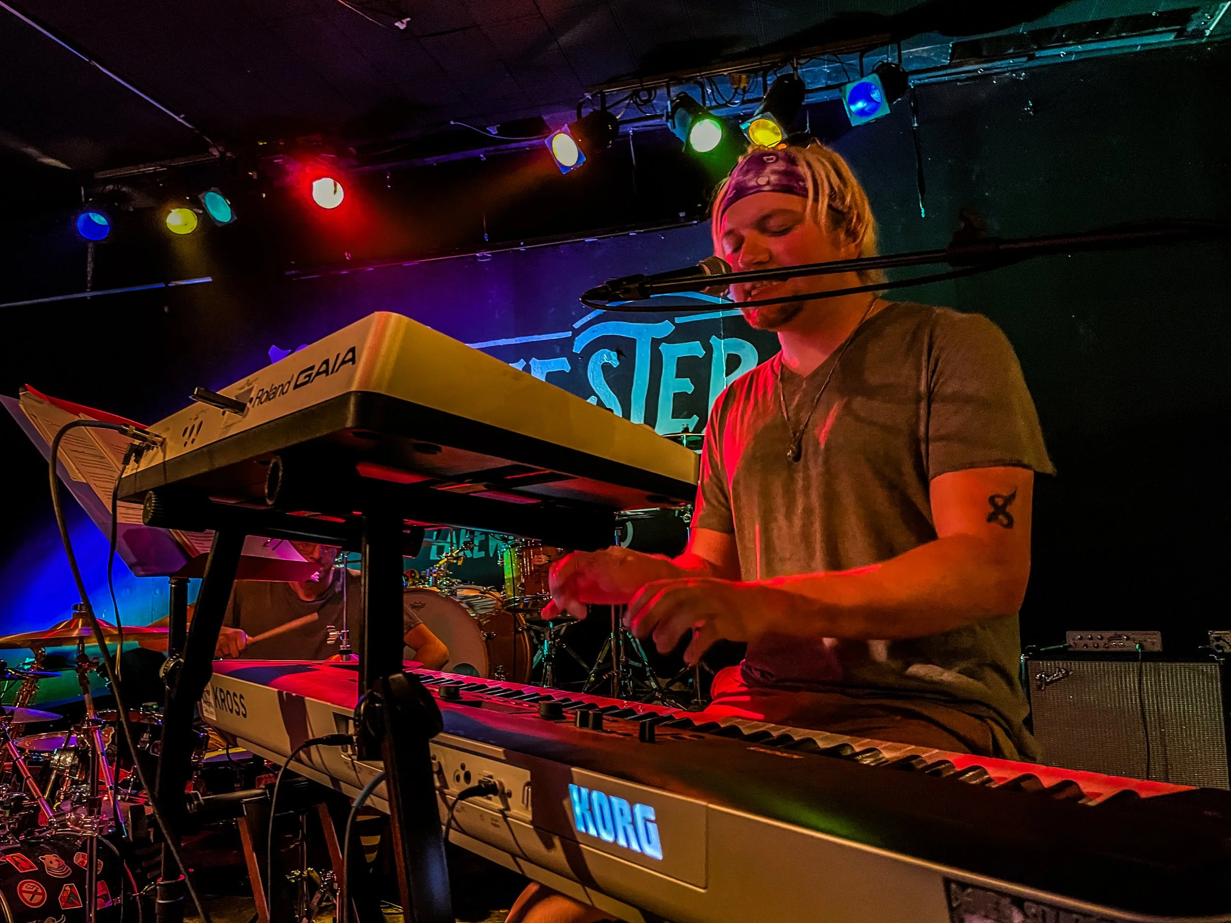 A musician with blond hair, a headband, and a tattoo on his arm, plays a keyboard on stage illuminated by colorful lights during a live performance.