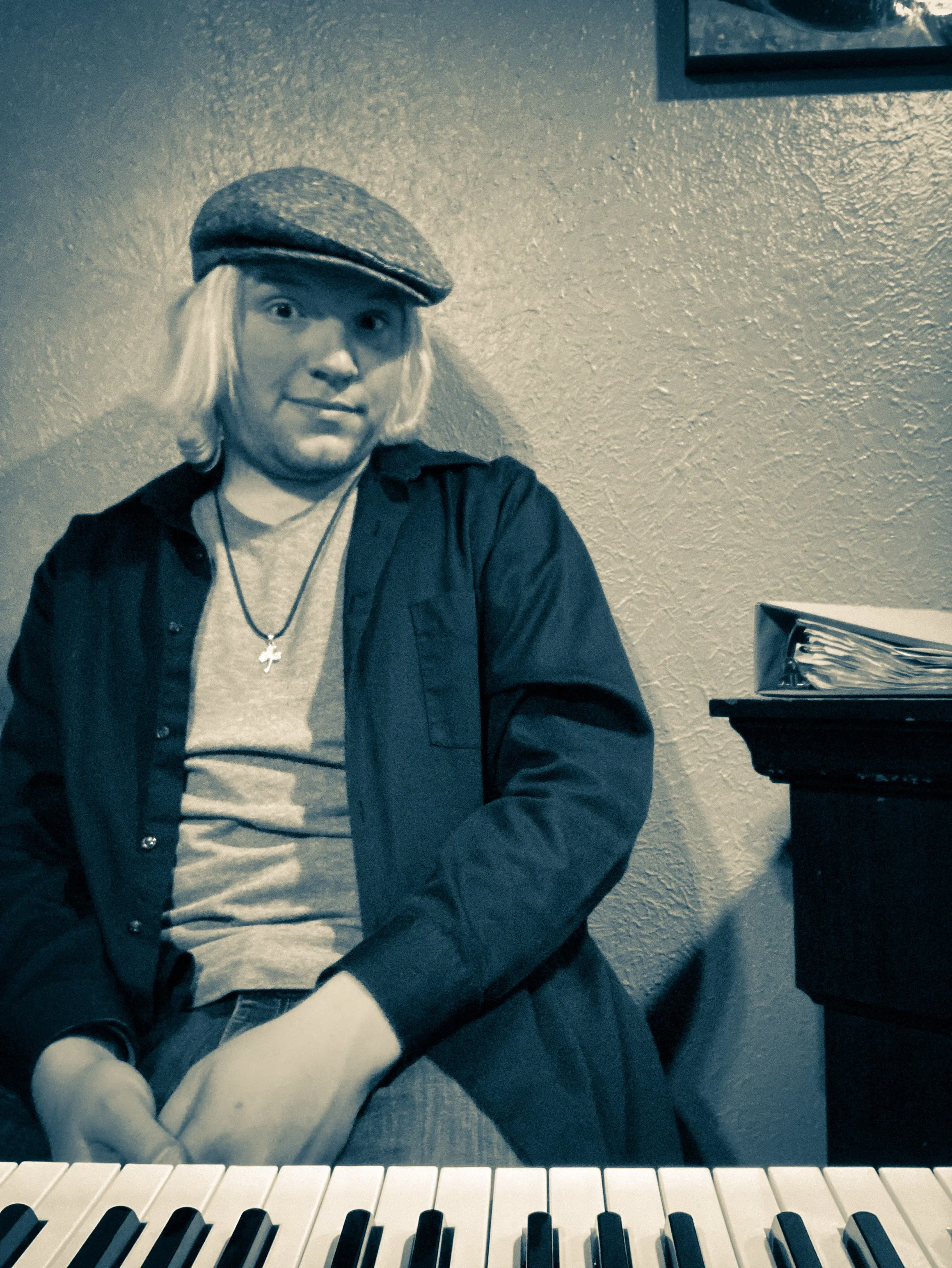 A person with shoulder-length blonde hair wearing a flat cap, a dark shirt, a light-colored t-shirt, and a necklace sitting in front of a piano.