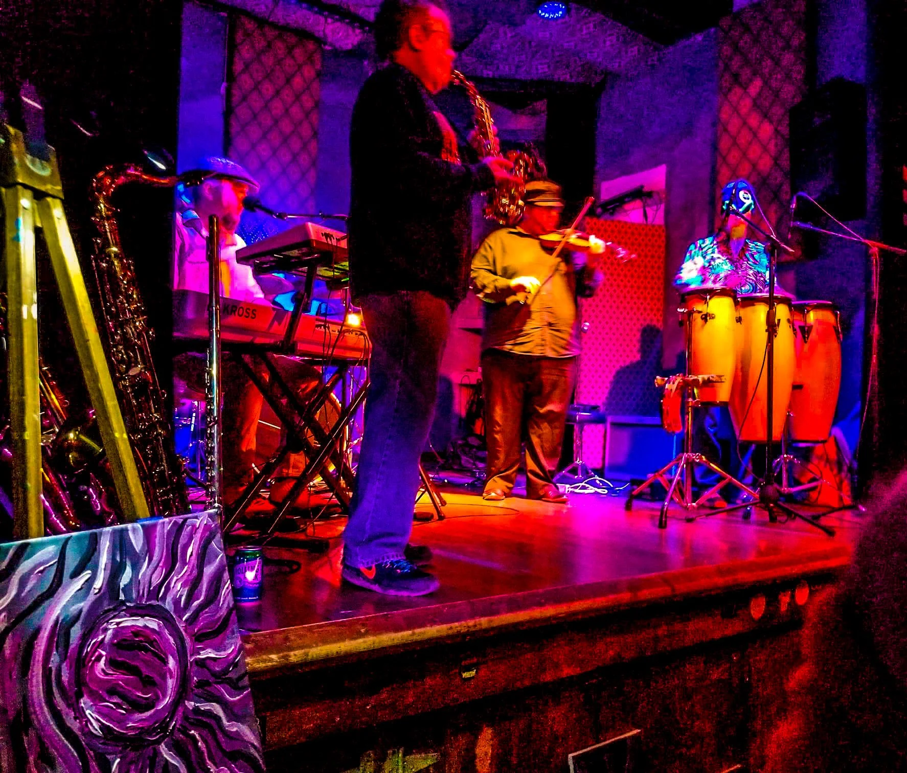 Musicians performing on a small stage with colorful lighting, including a saxophonist, keyboardist, violinist, and percussionists, in a cozy venue.