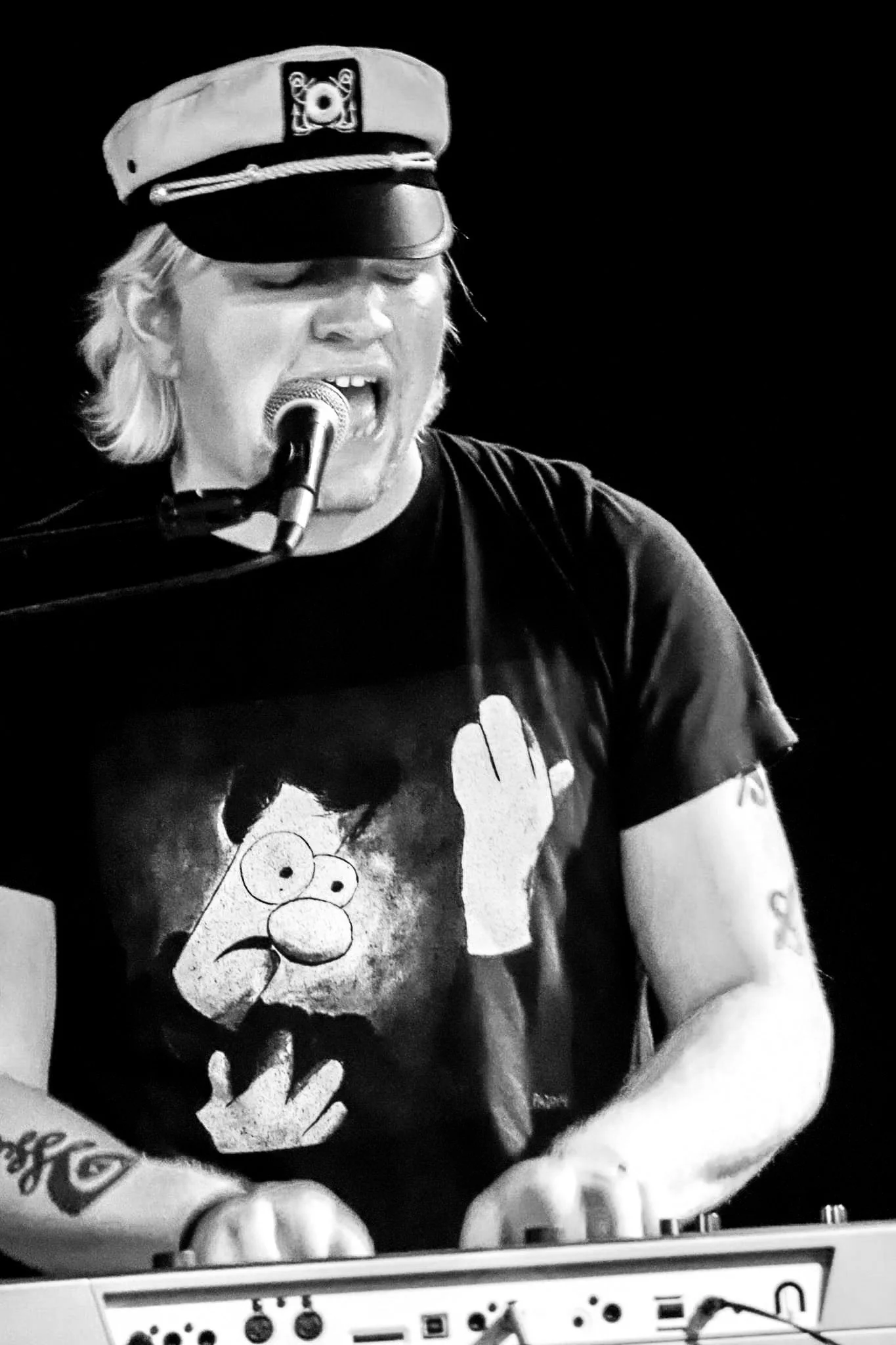 A man with long, light-colored hair wearing a captain's hat and a black t-shirt with cartoon characters, playing a keyboard and singing into a microphone.