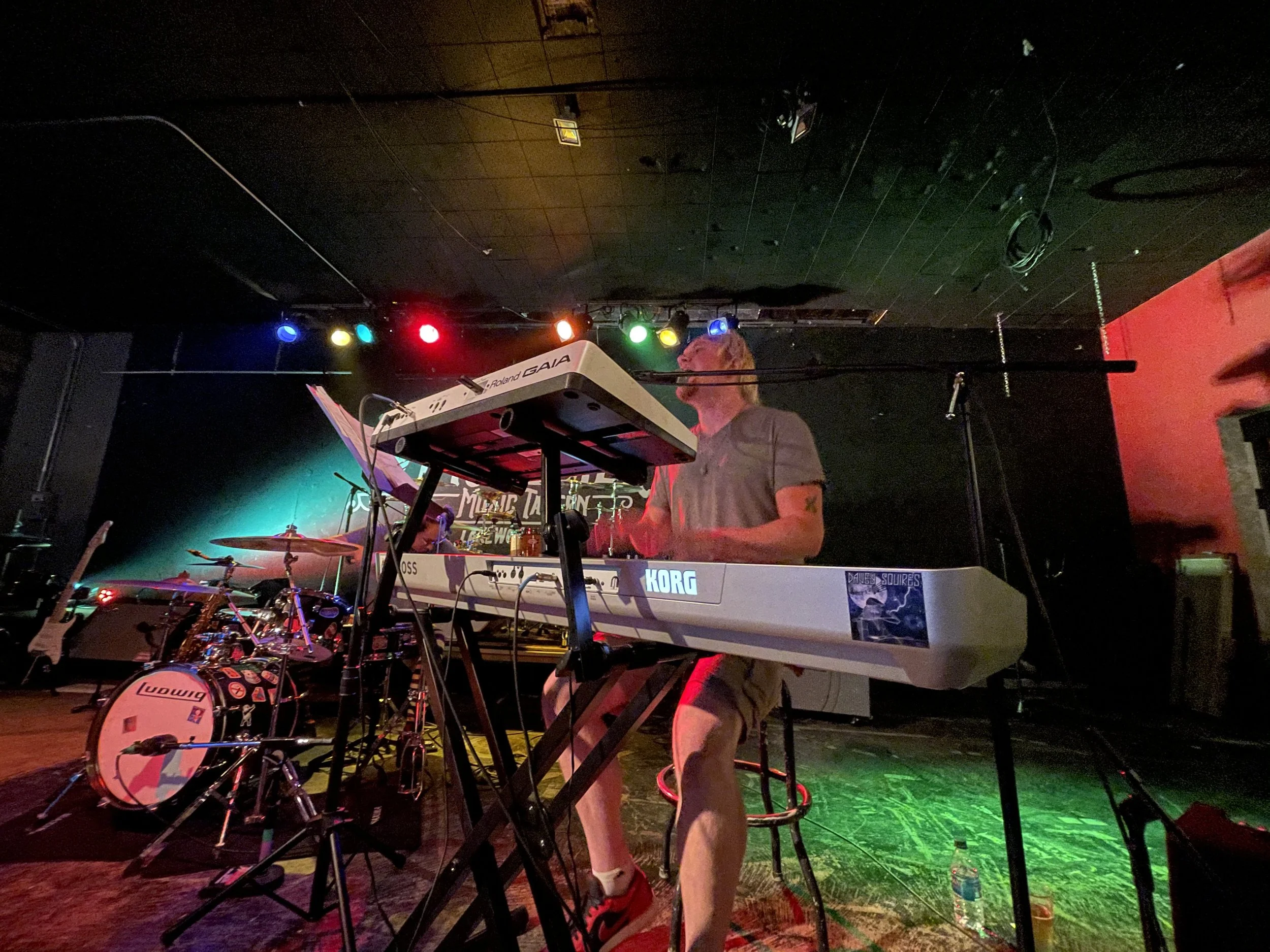 A musician playing a keyboard on stage with colorful stage lights, drums, and musical equipment around.