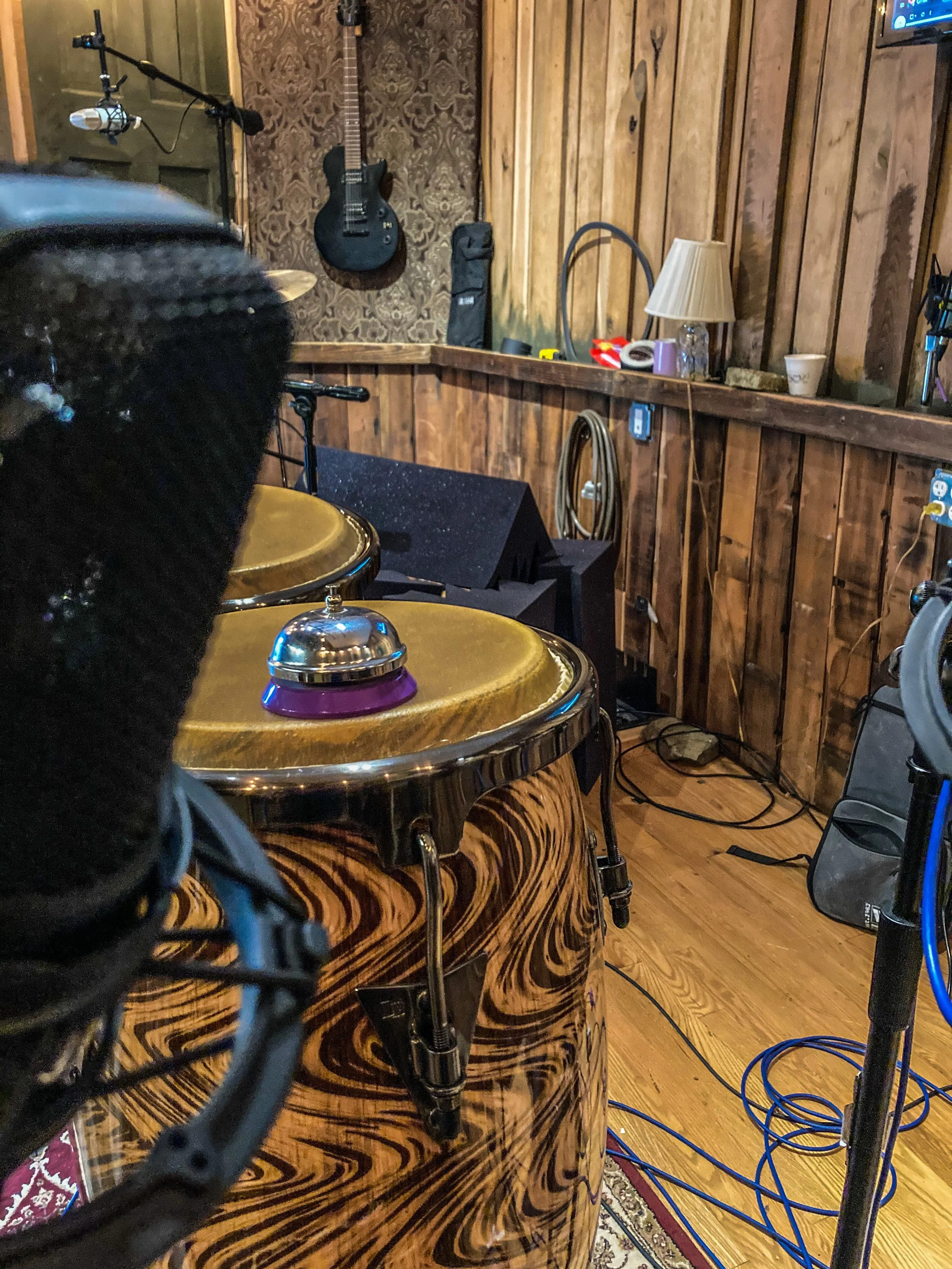 A music recording studio with wooden walls, featuring a guitar hanging on the wall, a lamp, and various audio equipment. The foreground shows a close-up of a percussion instrument with a metallic bell on top.