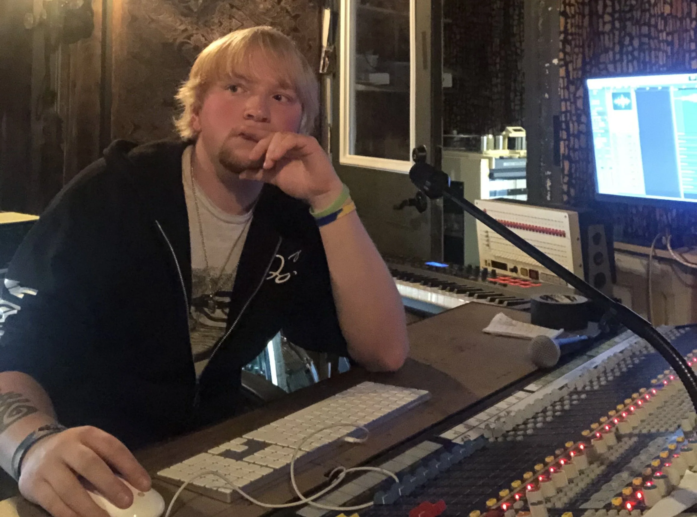 A young man with blond hair and a beard sitting in a recording studio, surrounded by audio equipment, a keyboard, computer screens, and a microphone, with his hand on a computer mouse.