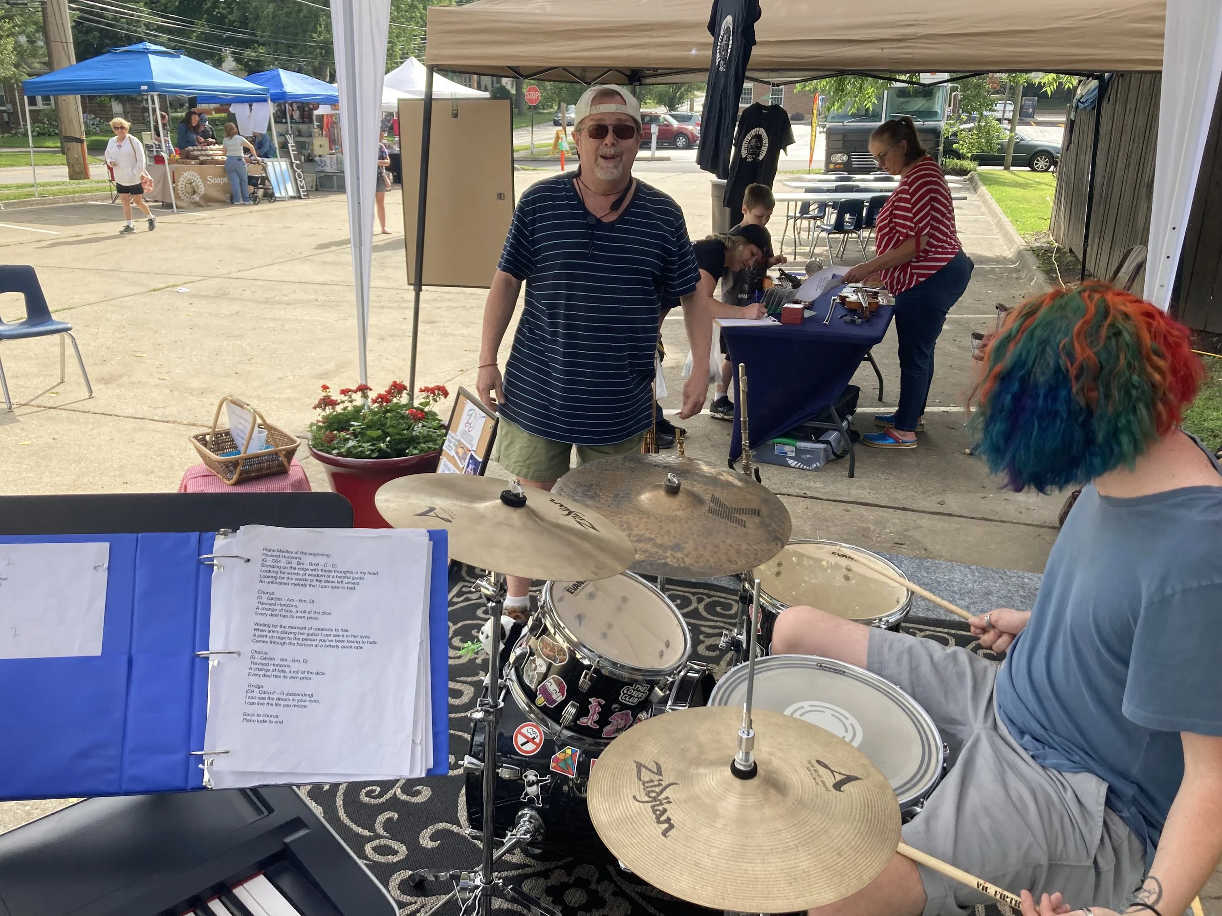 A man with rainbow-colored hair playing drums at an outdoor event with tents, vendors, and people in the background.