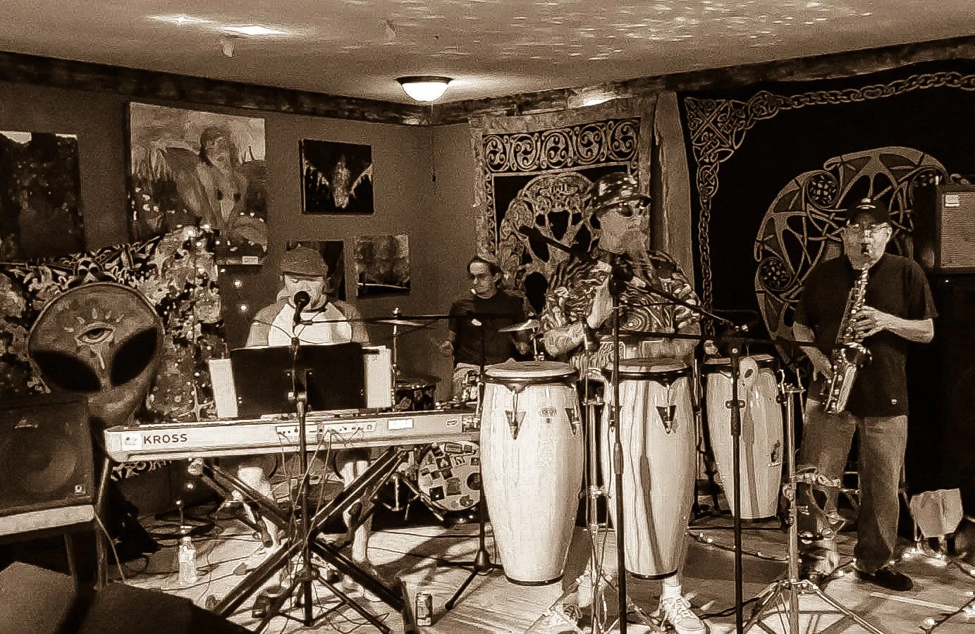 A band performing live on a small stage, featuring a keyboardist, percussionist, saxophonist, and a vocalist. The background is decorated with various artworks, and musical instruments are visible on stage.