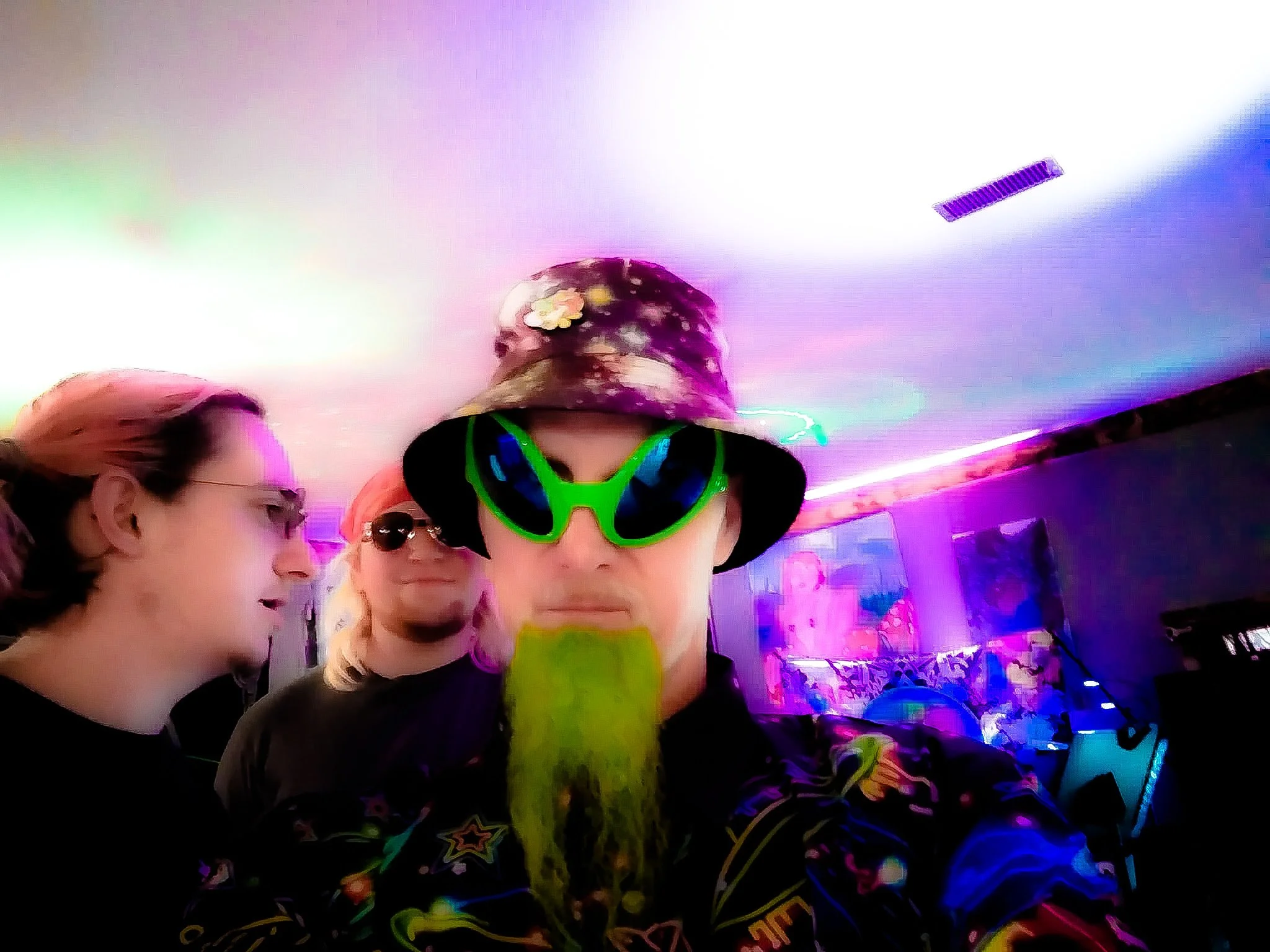 People at a party with colorful lighting, one wearing neon green sunglasses and a floral bucket hat, and others in dark clothing and sunglasses.