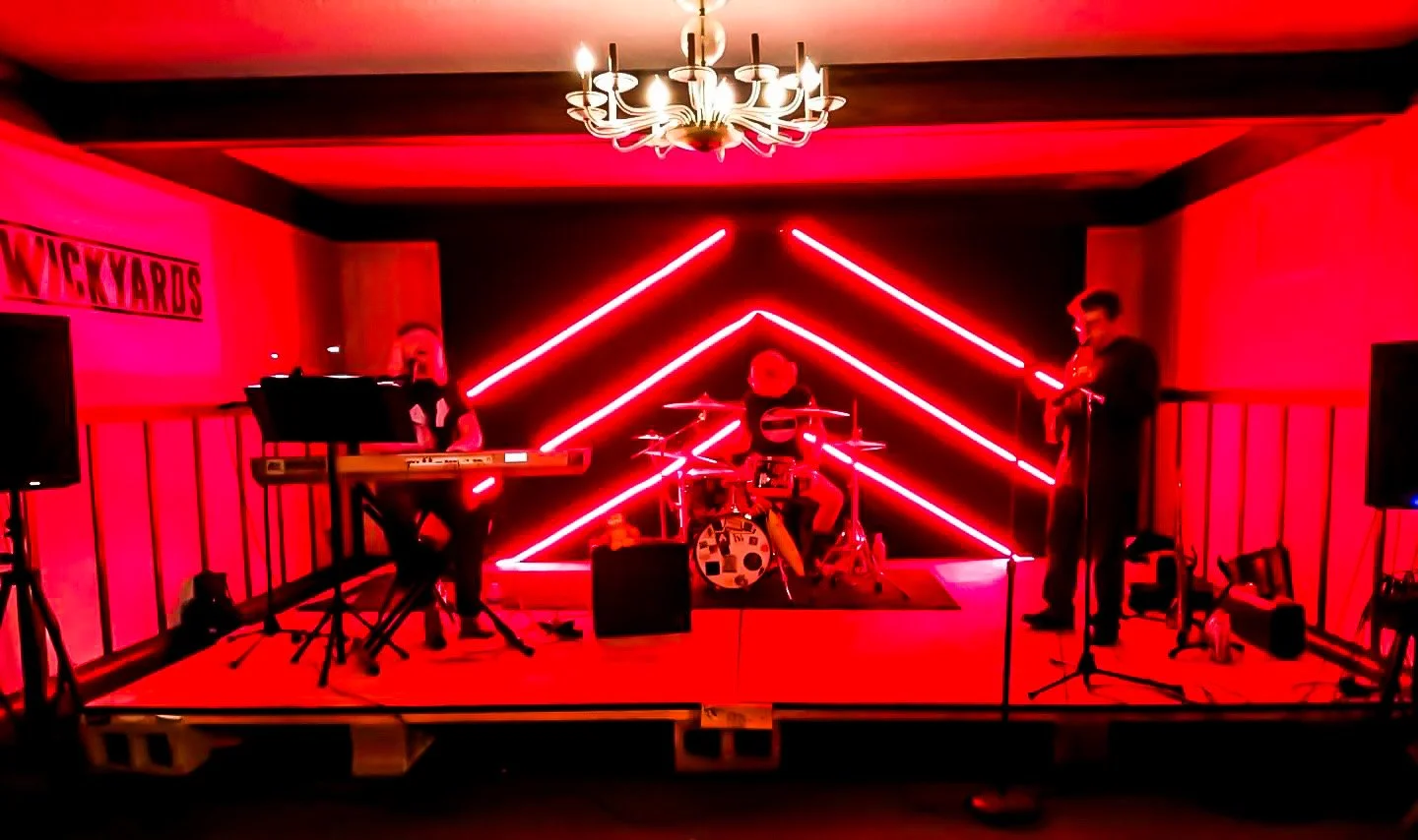 Band performing on stage with red neon lights in background, keyboard player on left, drummer in center, violinist on right, in a decorated room with chandelier.