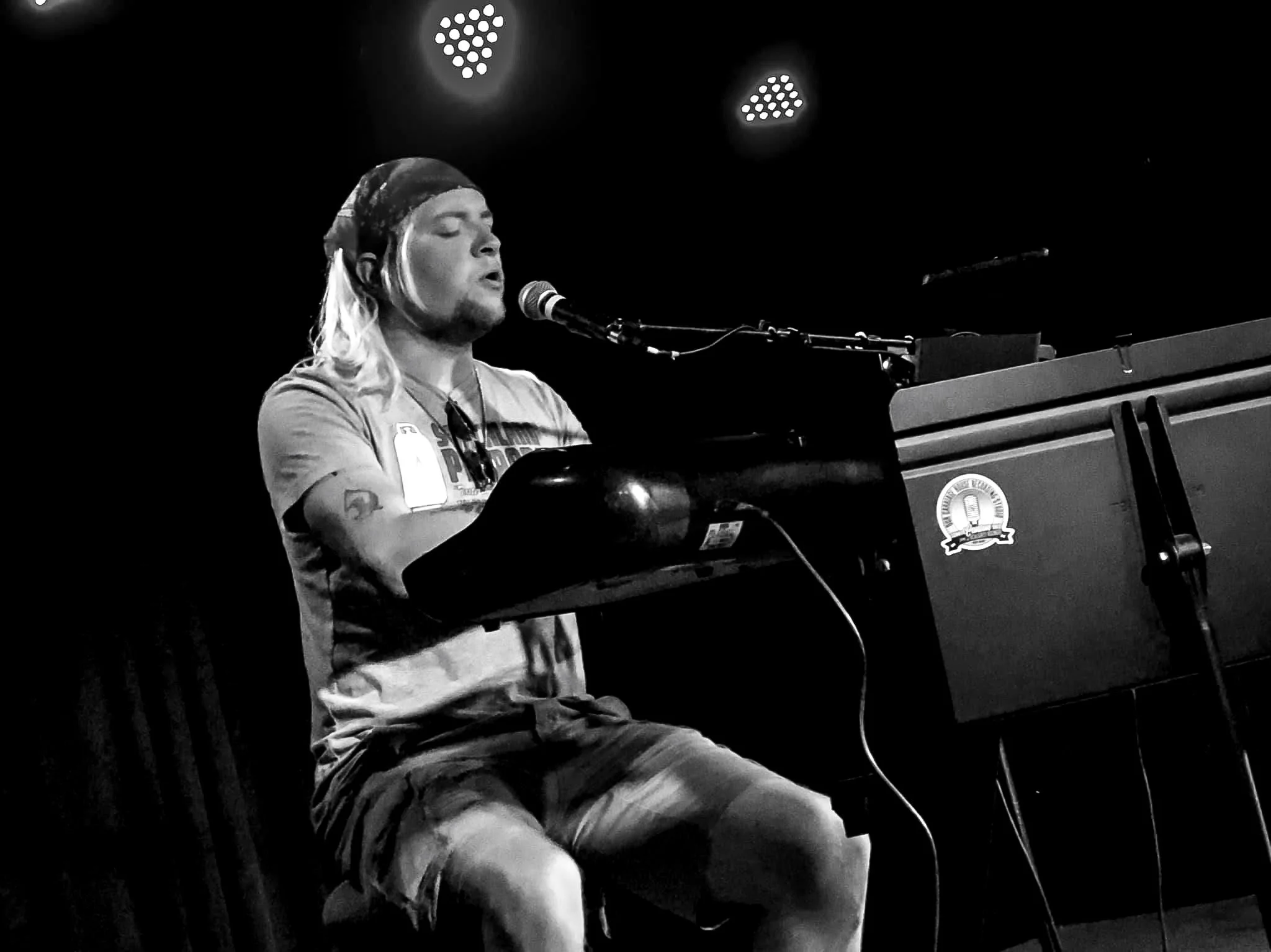 A man with long hair, a beard, and a bandana, singing into a microphone while playing a piano on stage, with two bright stage lights overhead.