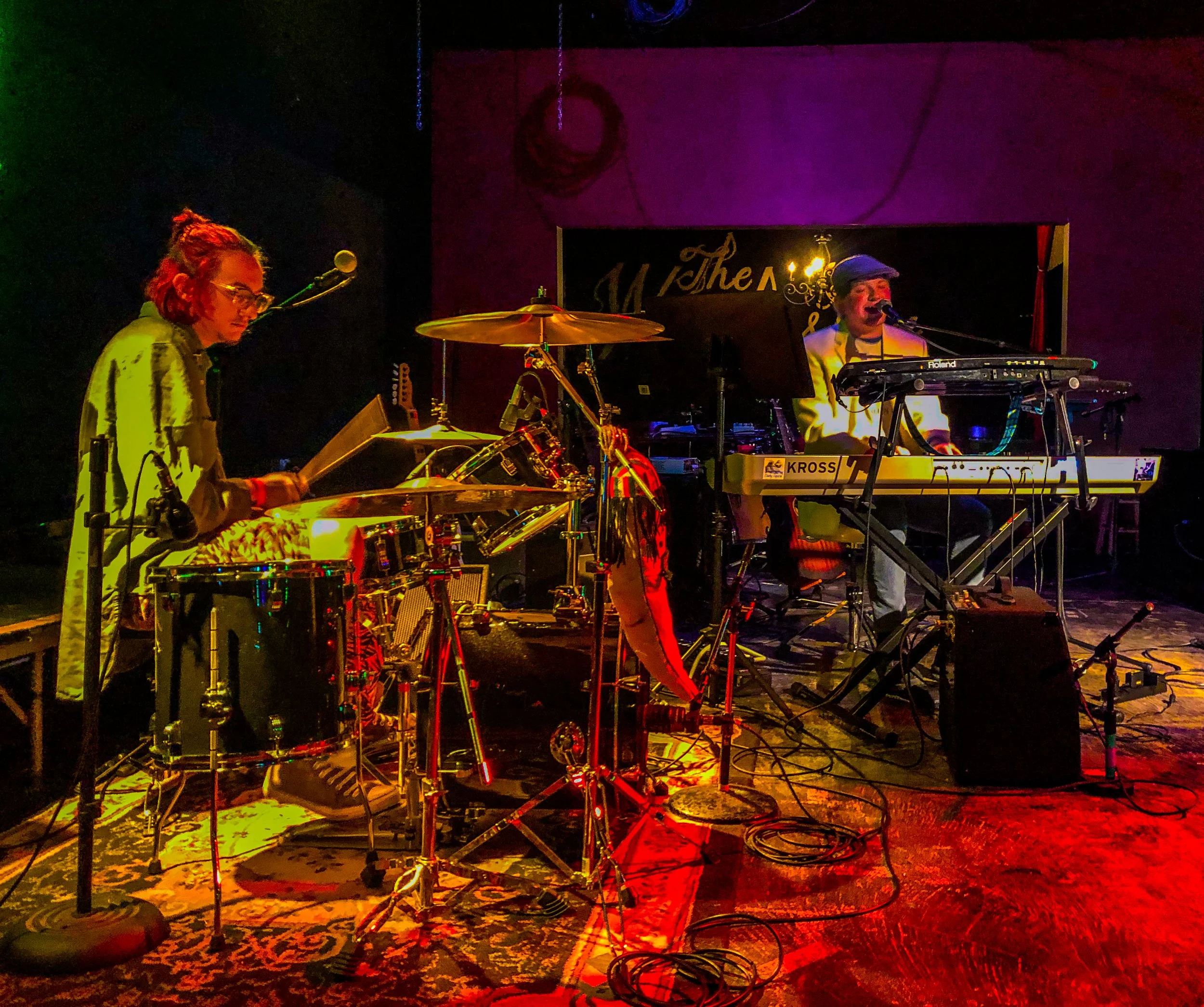 Musicians performing on stage at a concert, with a woman playing drums and a man singing and playing keyboard, illuminated by colorful stage lights.