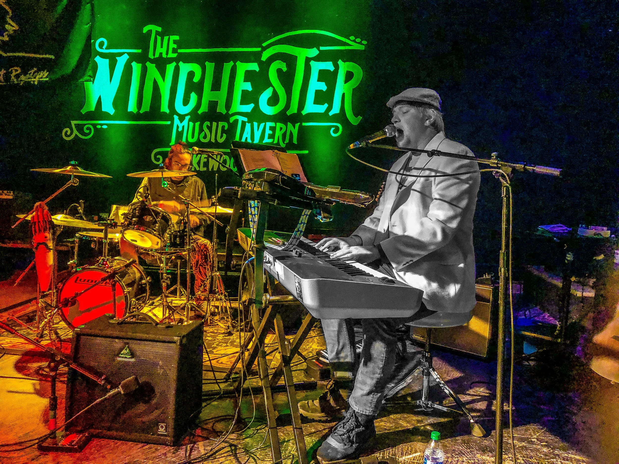 A band performing live at Winchester Music Tavern, with a keyboard player singing into a microphone and a drummer playing drums behind him. The stage is dimly lit with colorful lights and a bright green sign in the background reading 'The Winchester