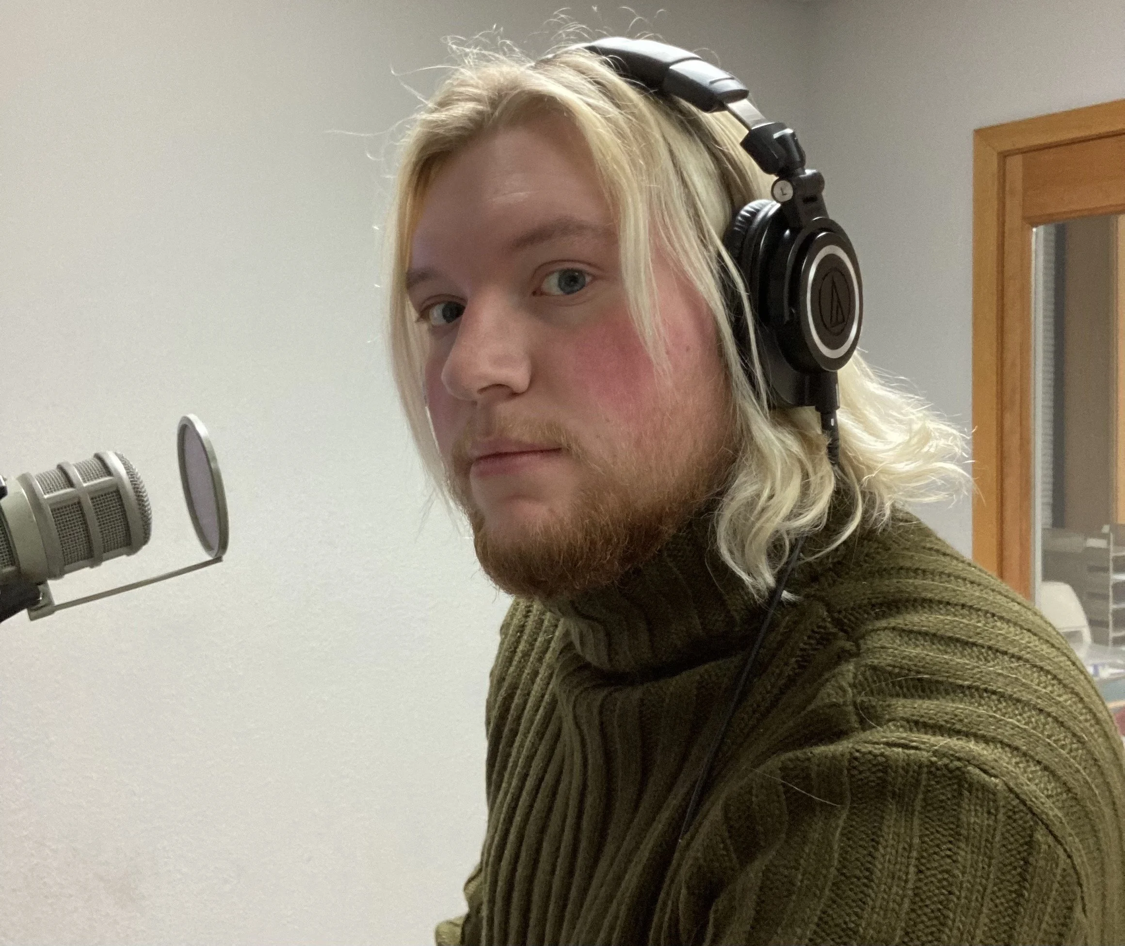A young man with long blond hair, headphones, and a beard sits in a recording studio near a microphone.